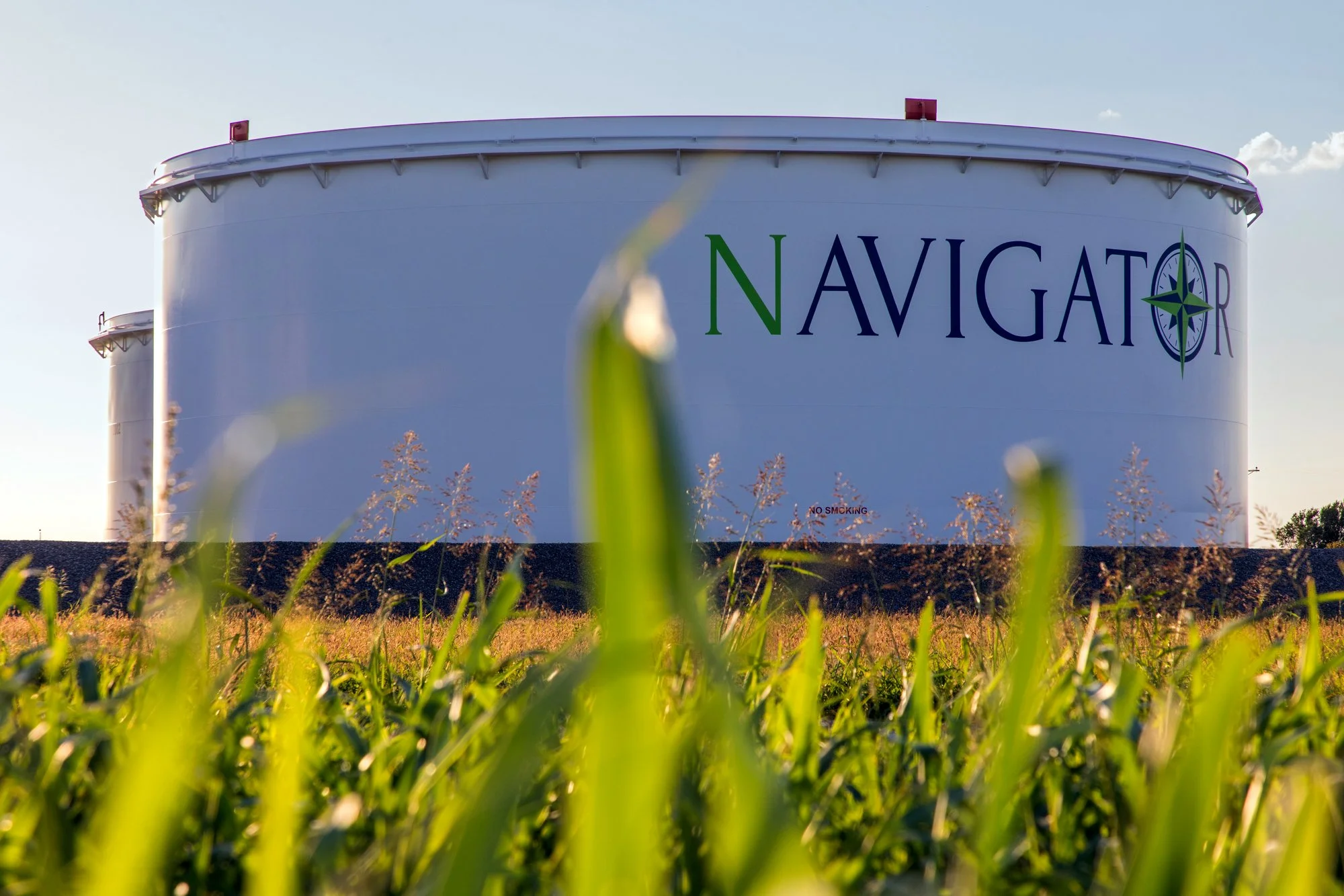 Large white water storage tank with the word NAVIGATOR in large colorful letters and a compass logo, seen from low angle in a grassy field during daytime.