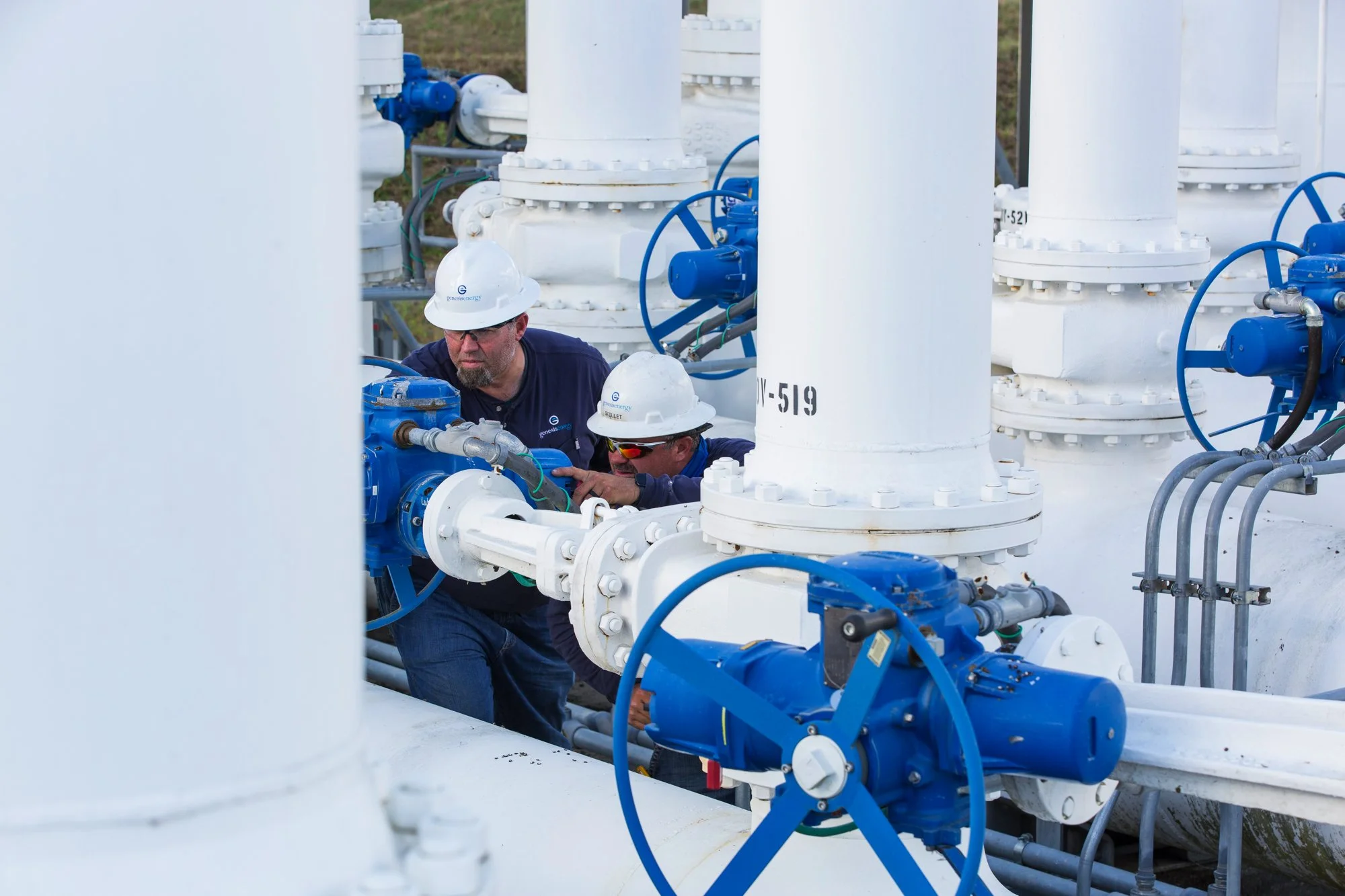 Two workers wearing white safety helmets and blue uniforms working on industrial pipes with blue valves and fittings.