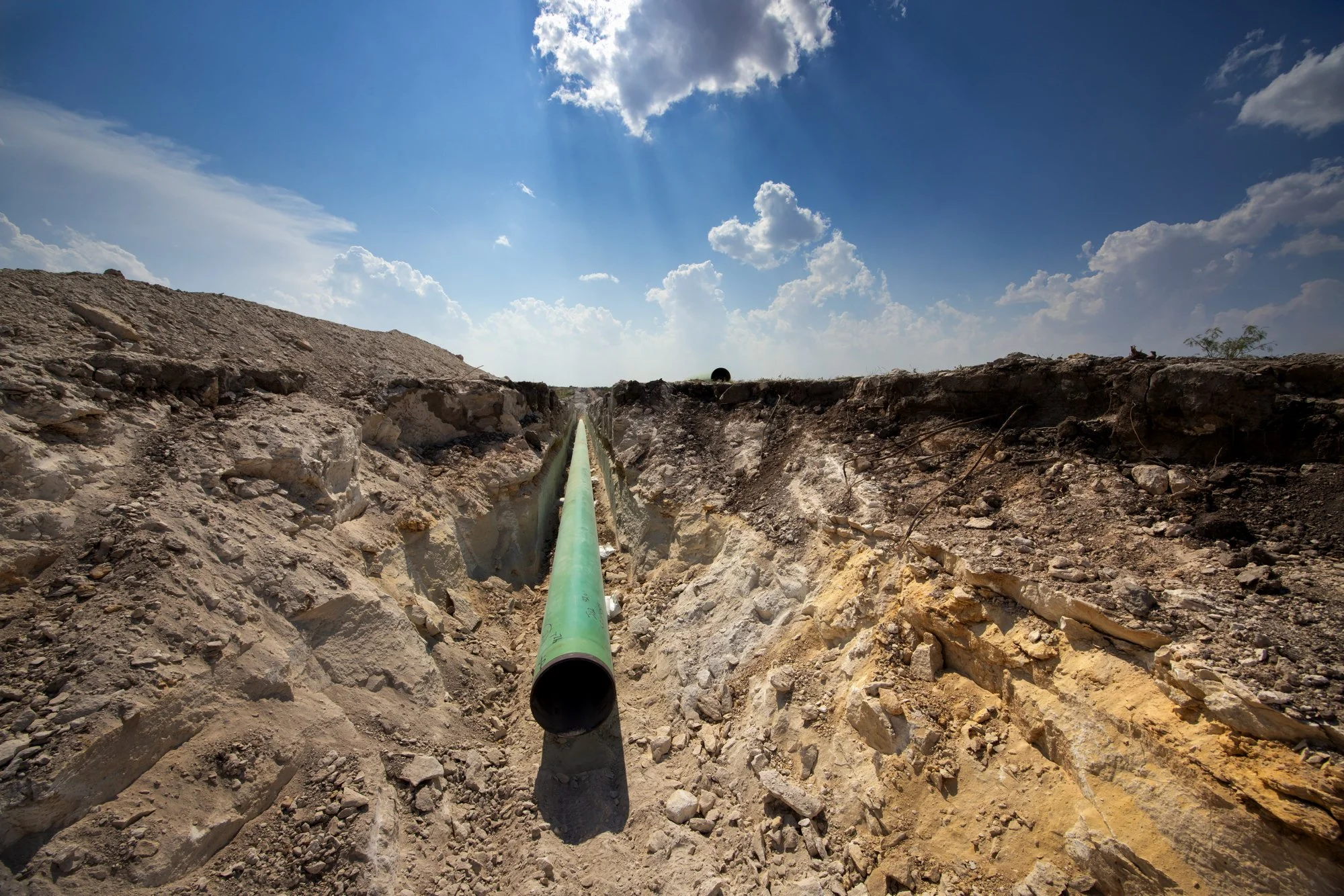 A green pipeline running through a trench in a dry, rocky landscape under a partly cloudy sky.