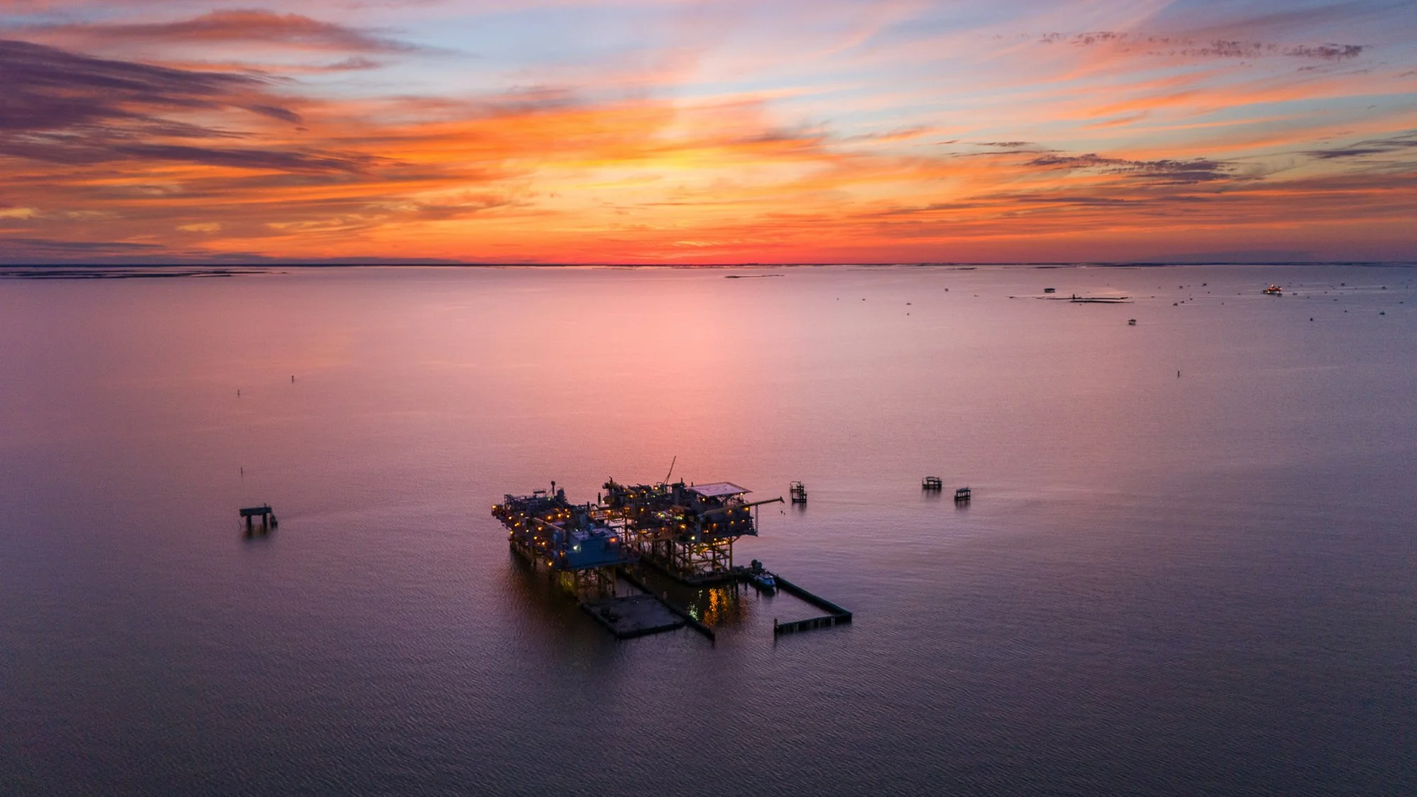 Aerial view of an offshore oil platform at sunset over calm ocean water, with a colorful sky and distant boats and structures.