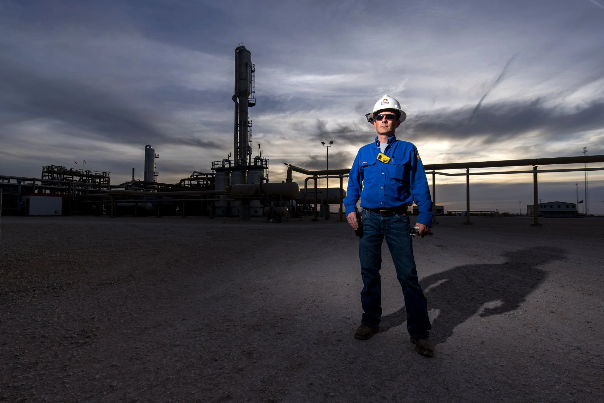 A man in safety gear stands in front of an industrial plant with pipes and smokestacks during cloudy weather at sunset.