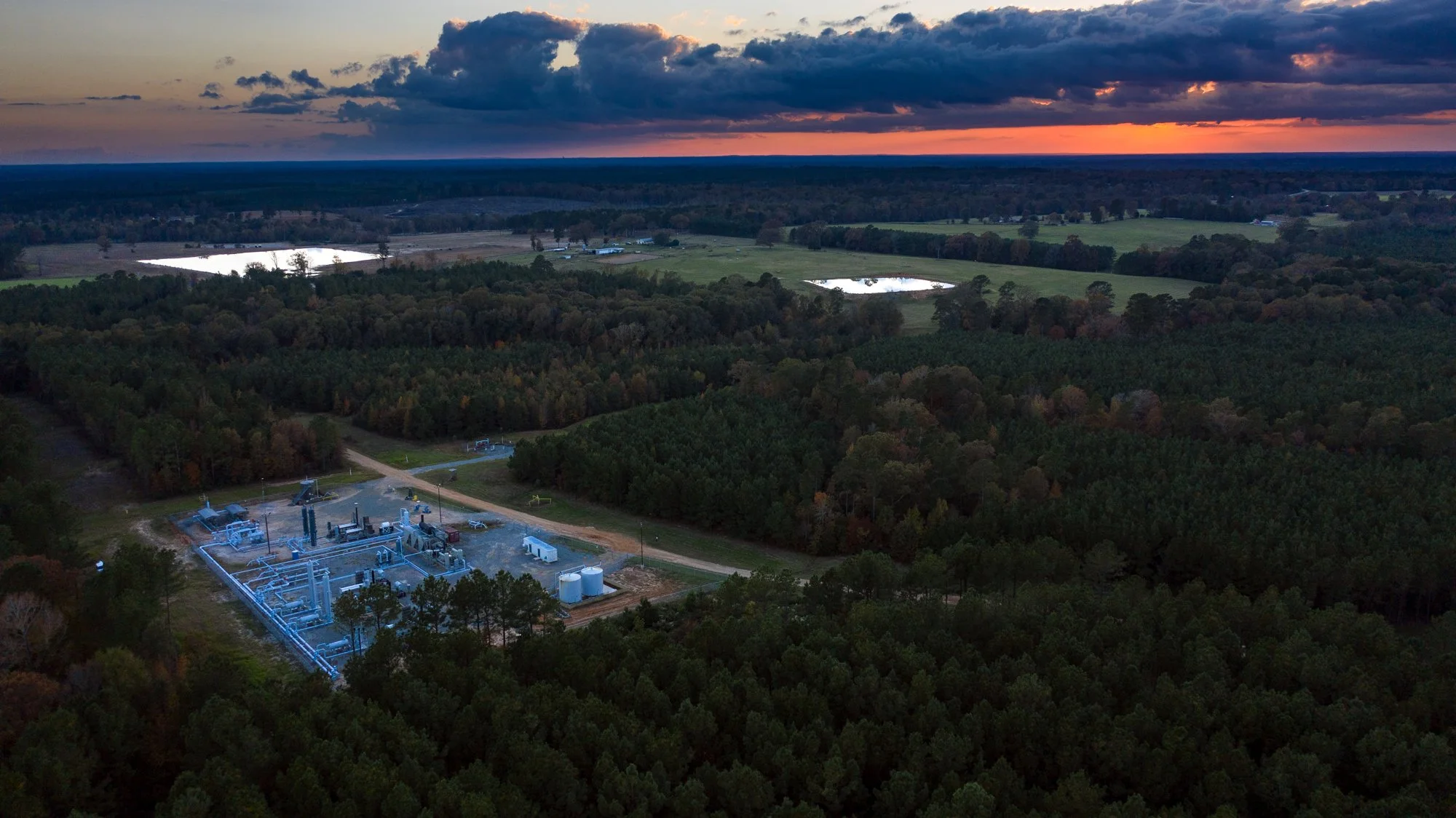 Aerial view of a large industrial facility surrounded by dense forests, with ponds and fields in the background during sunset.