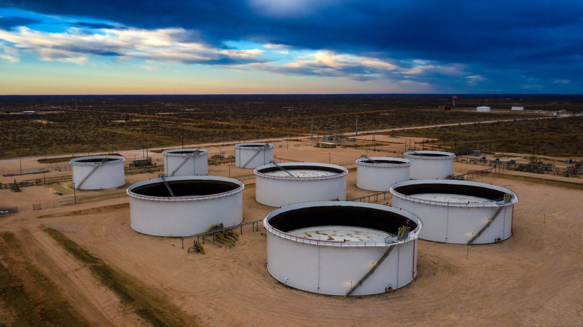 Aerial view of large industrial oil storage tanks in a desert landscape during sunset, with a dirt road nearby and a vast flat terrain extending to the horizon under a partly cloudy sky.