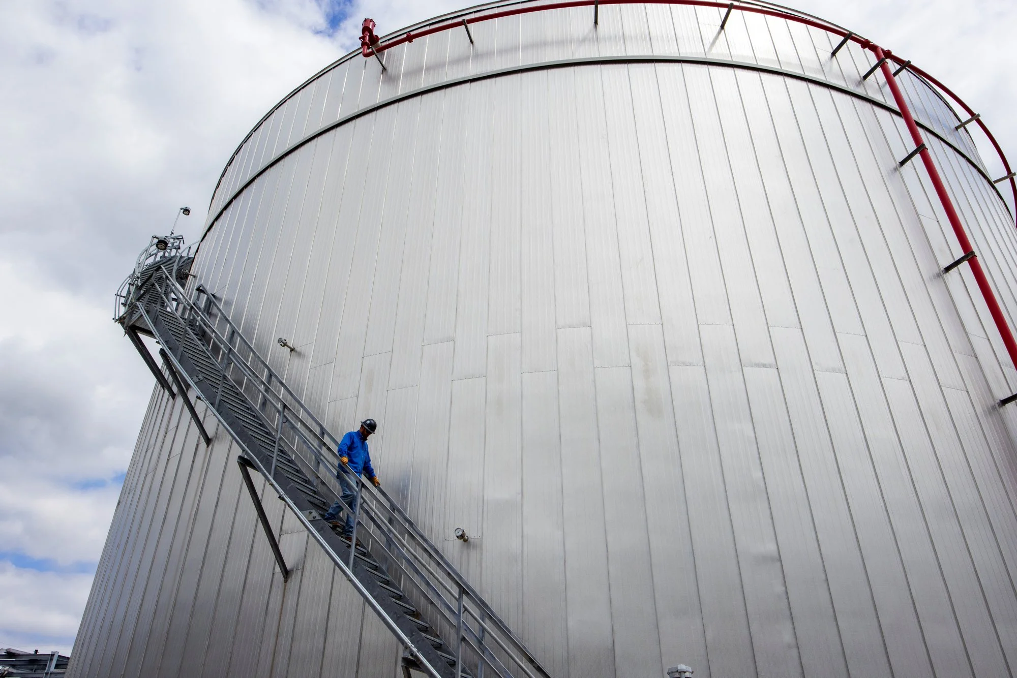 A worker in blue safety gear and a helmet walking down a metal staircase outside a large cylindrical storage tank with a cloudy sky in the background.