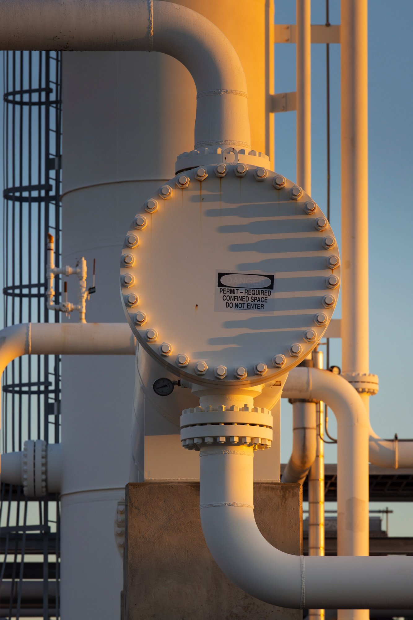Industrial piping system with large white valve, pipes, and a safety sign that reads 'Permit-Required Confined Space Do Not Enter' at sunset.