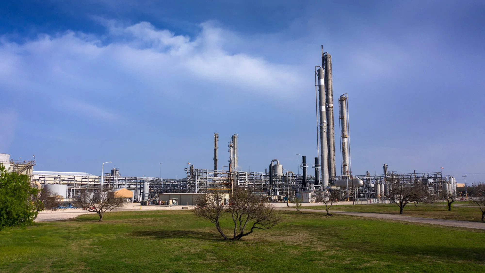 Industrial facility with multiple tall pipes and storage tanks, surrounded by a grassy area with sparse trees and a clear blue sky.