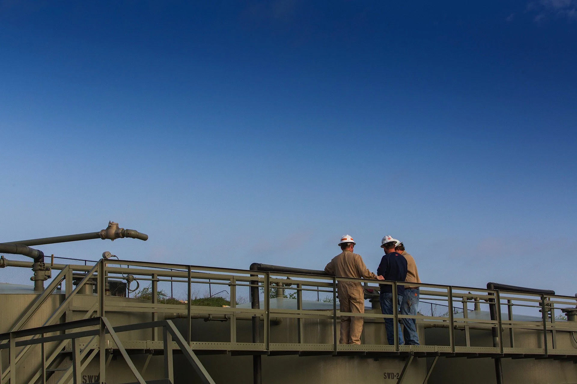 Three workers in safety helmets working on a large industrial tank under a clear blue sky.