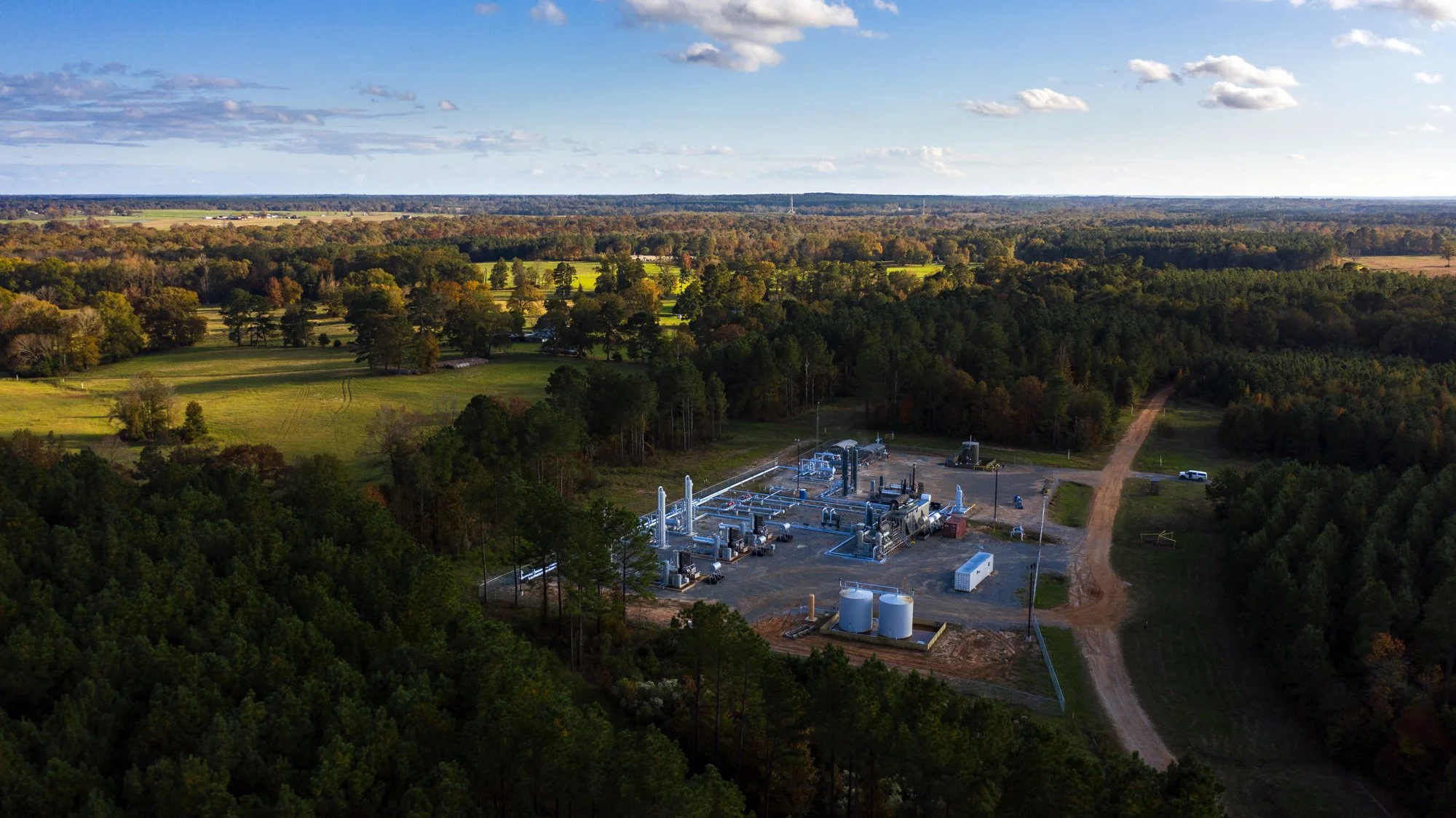 Aerial view of an industrial facility surrounded by green trees and open fields with a dirt road leading to it.