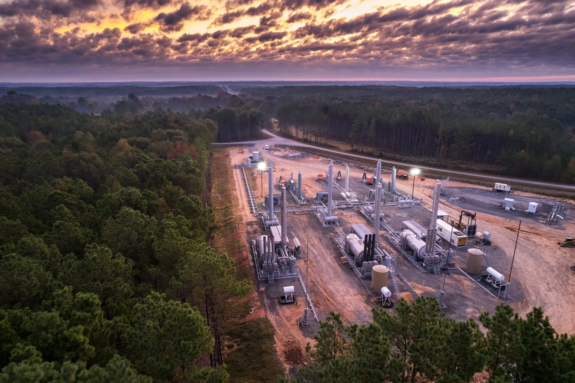Aerial view of a natural gas processing plant located in a forested area with a dirt road nearby during sunset.