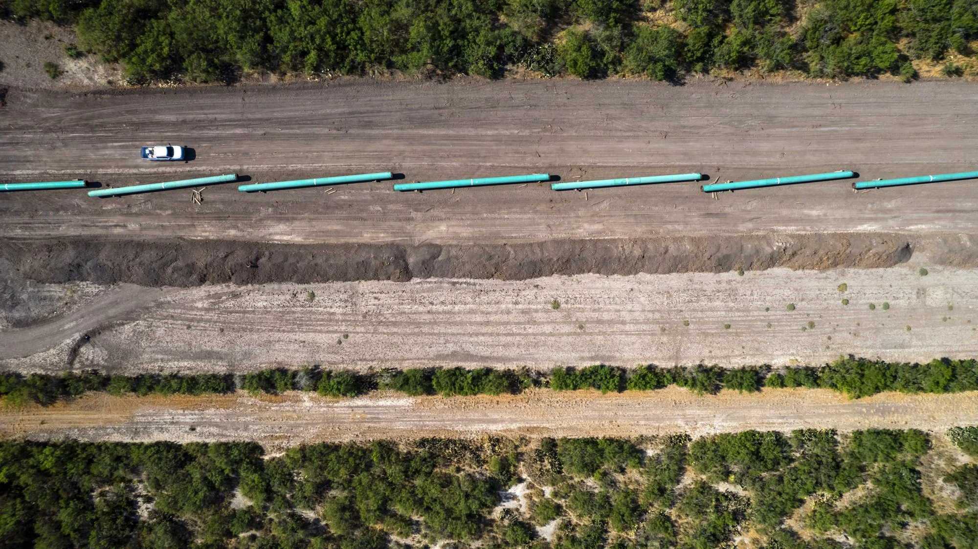 Aerial view of a pipeline construction site with large blue pipes laid out on the ground, surrounded by trees and dirt roads.