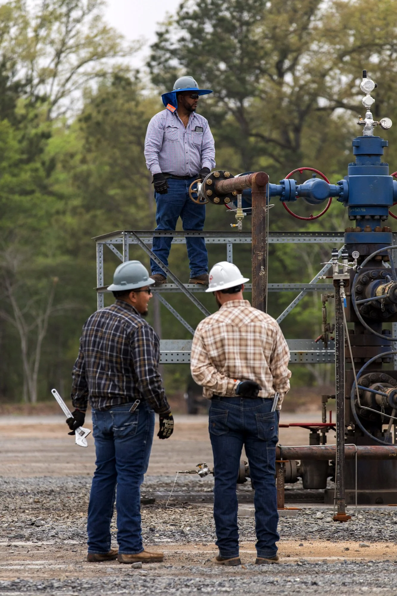 Three workers wearing hard hats and gloves working on industrial equipment outdoors. One worker stands on a metal platform adjusting a valve, while two others stand below observing and holding tools. The background has trees and a cloudy sky.