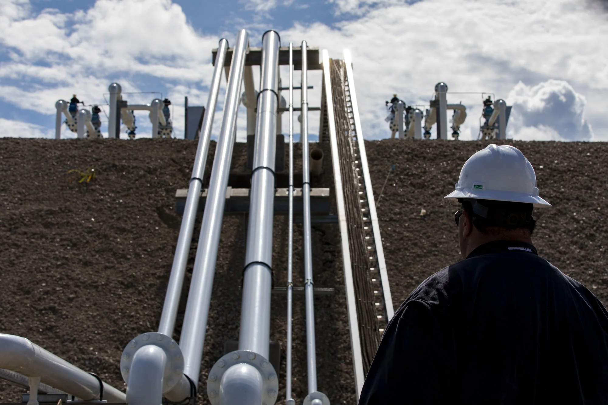 A man wearing a white safety helmet and black jacket is observing industrial pipes and equipment on a construction site against a cloudy sky.