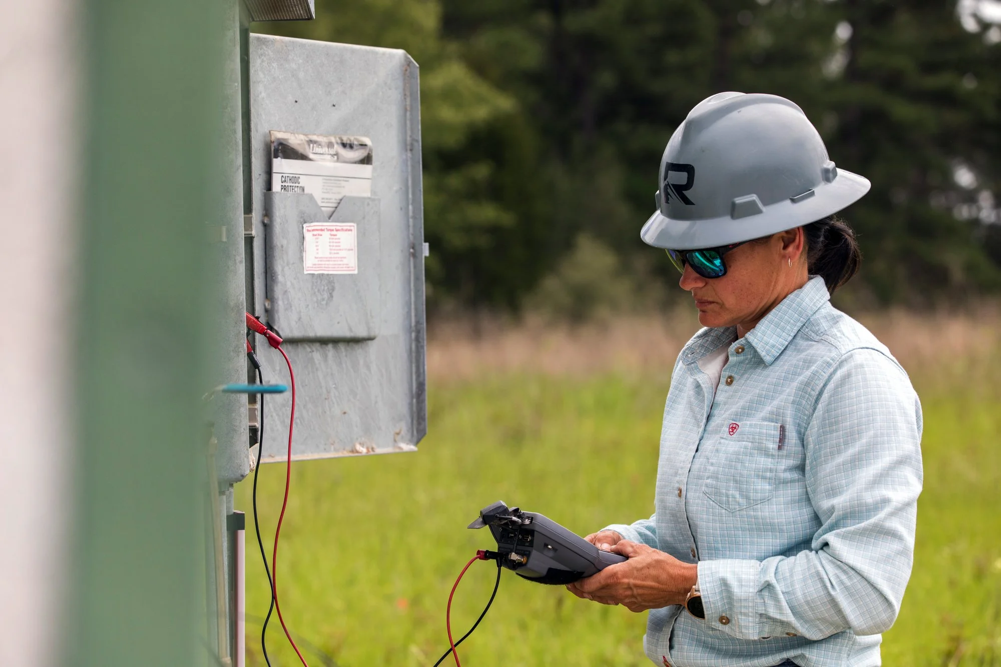 A female utility worker in a hard hat and sunglasses checking electrical equipment outside in a grassy area.