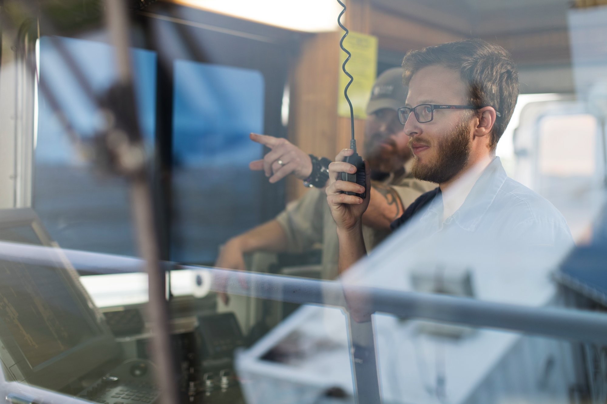 Two men working inside a control room with a glass window. One man is pointing at computer screens, and the other is holding a radio device.