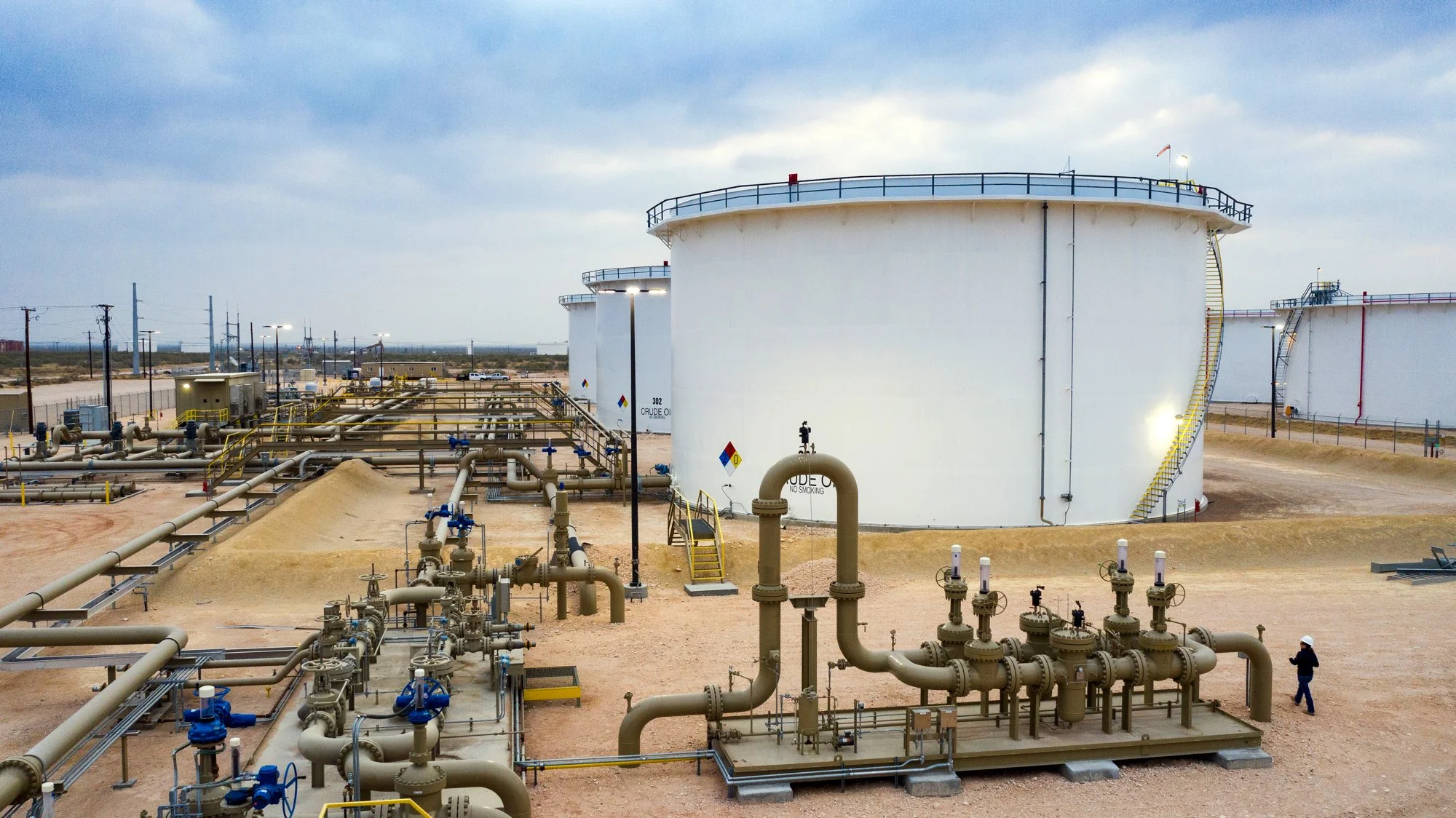 Oil storage tanks in an industrial oil field with pipelines, valves, and equipment, in a desert landscape under a cloudy sky.