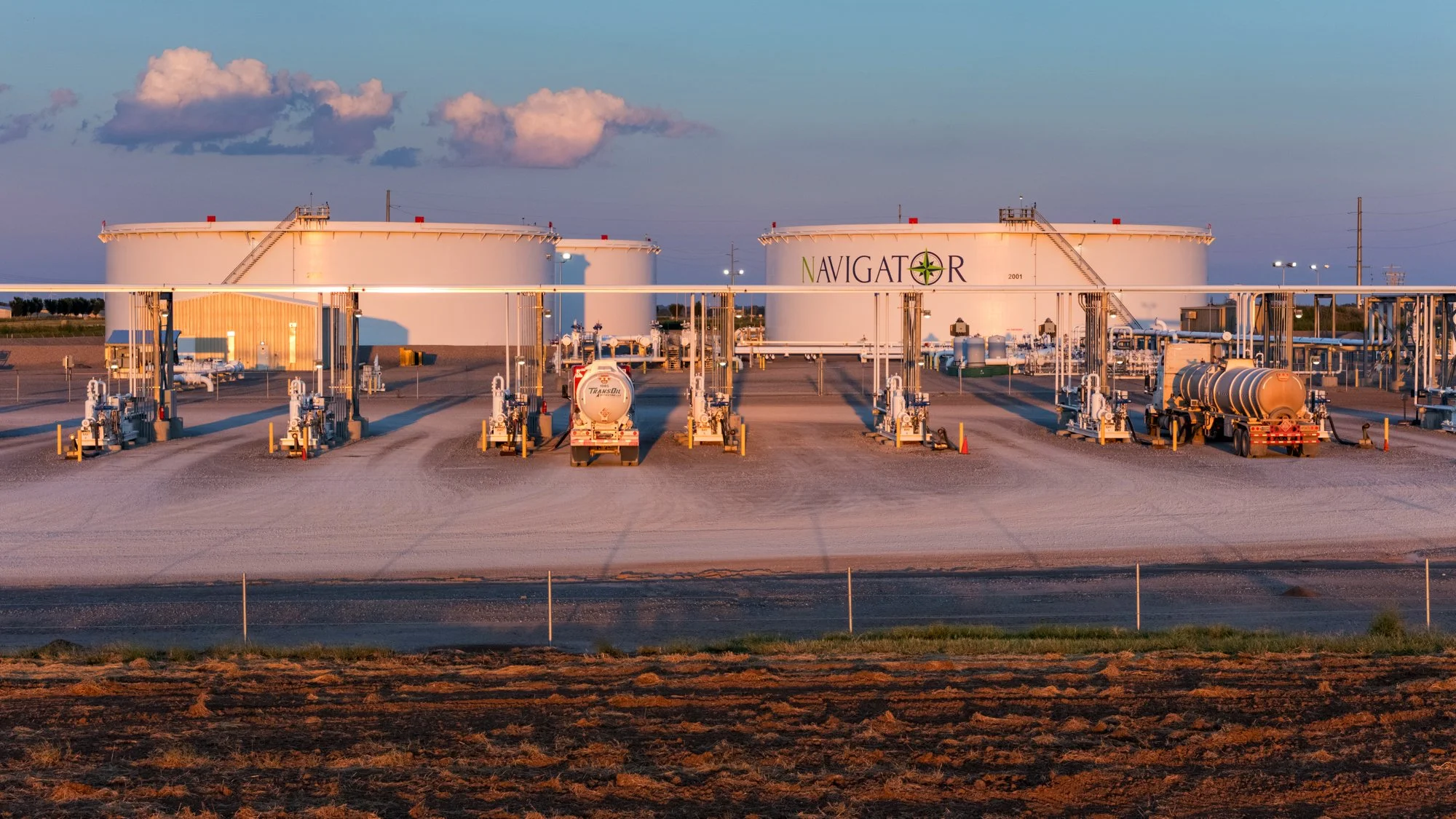 Oil storage tanks labeled 'Navigator' with fueling equipment and trucks in an oil depot at sunset.