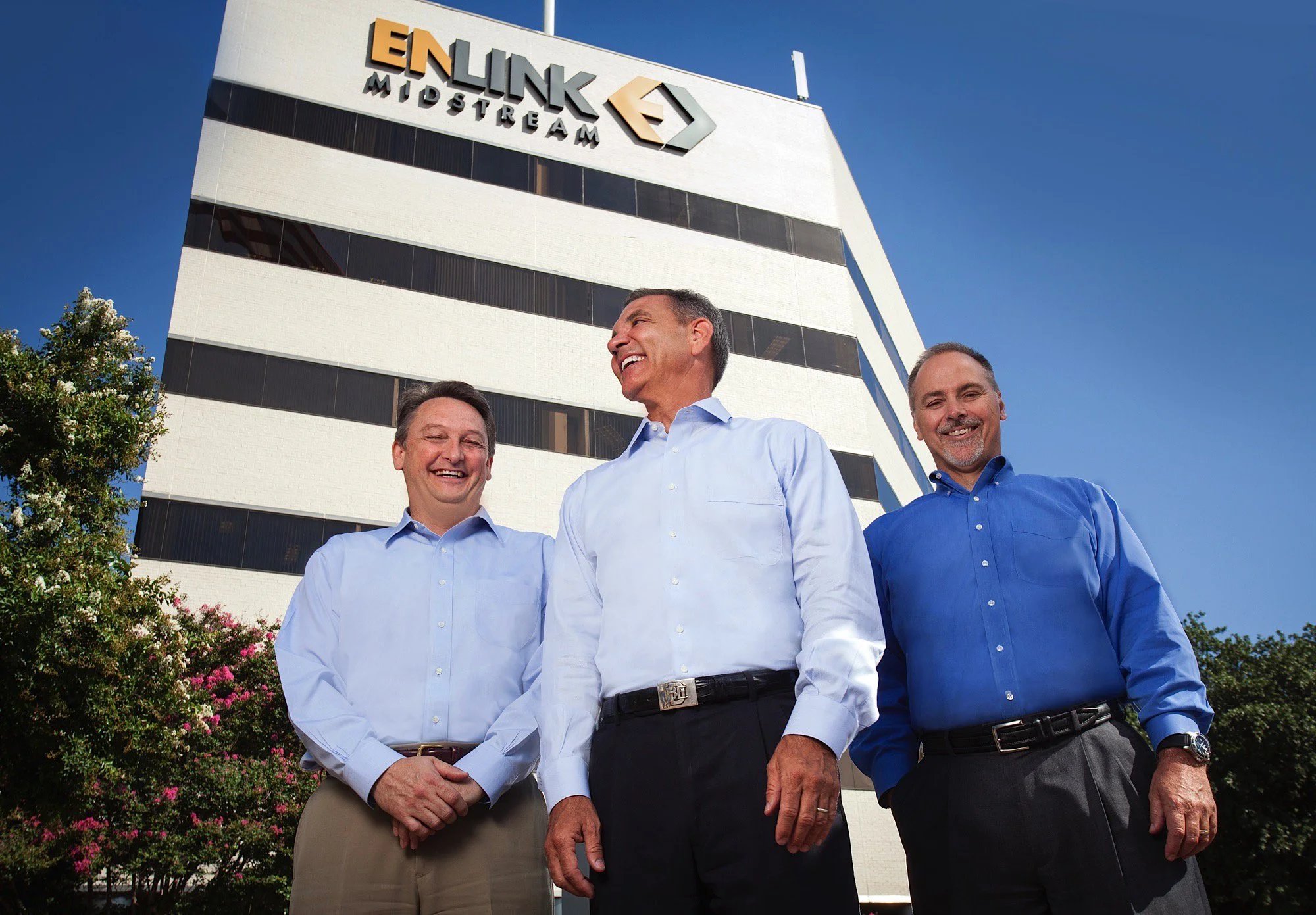 Three businessmen in dress shirts standing outdoors in front of a tall office building with the ENLINK Midstream logo, smiling and talking.