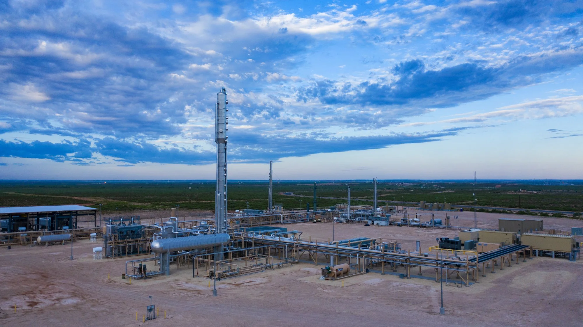 An oil or gas processing facility with pipelines and tall towers, set against a vast, flat landscape under a partly cloudy sky.