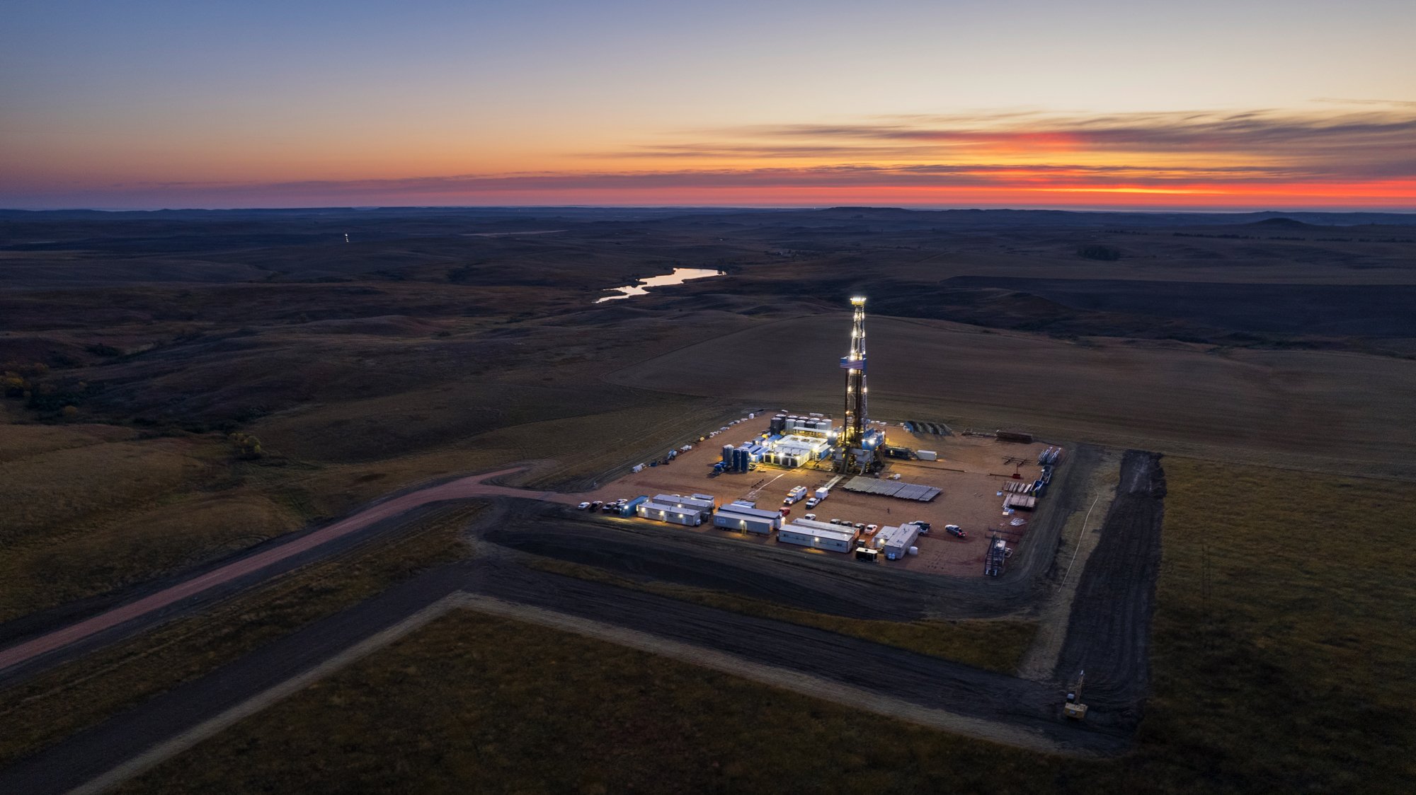 An aerial view of an oil drilling rig at sunset in a rural landscape with open fields and a small river.