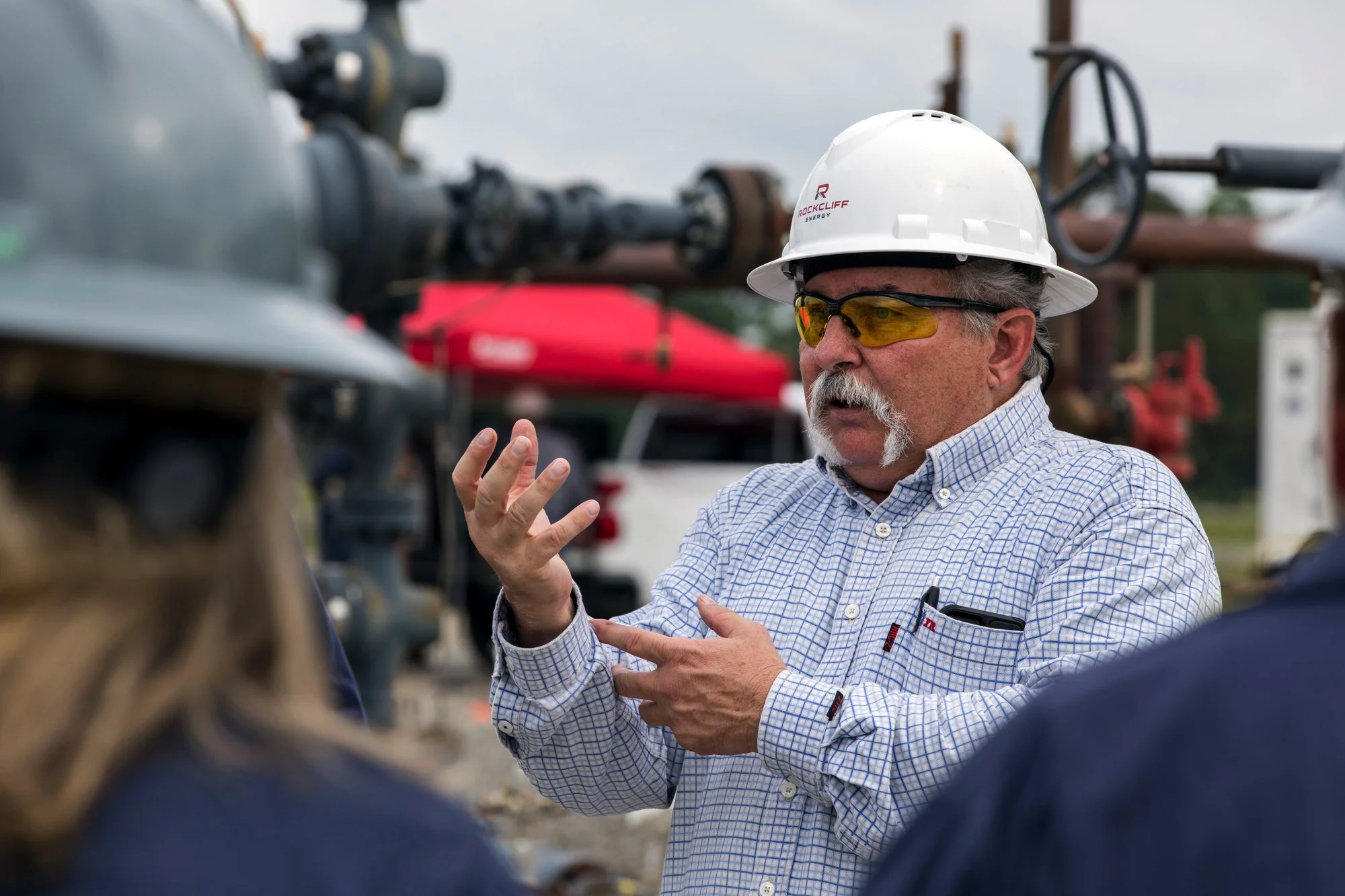 A man wearing a white hard hat, safety glasses, and a checkered shirt is explaining something to a group outside near industrial equipment. There are people around him, blurred in the foreground, and a red tent and a white vehicle are visible in the 
