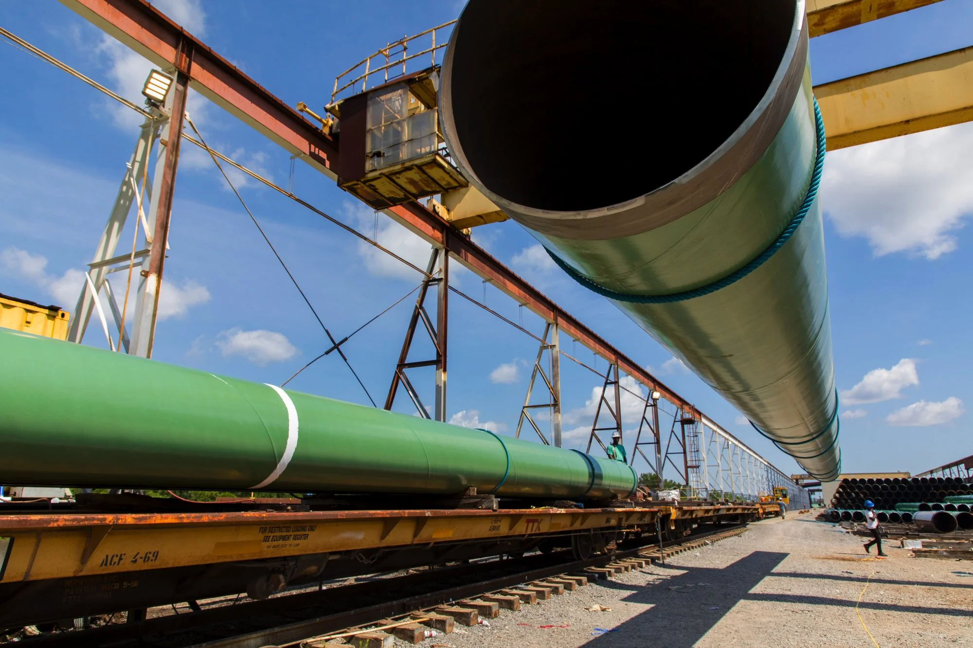 Large green pipe being transported on a flatbed railroad car at an industrial construction site under a blue sky with scattered clouds.