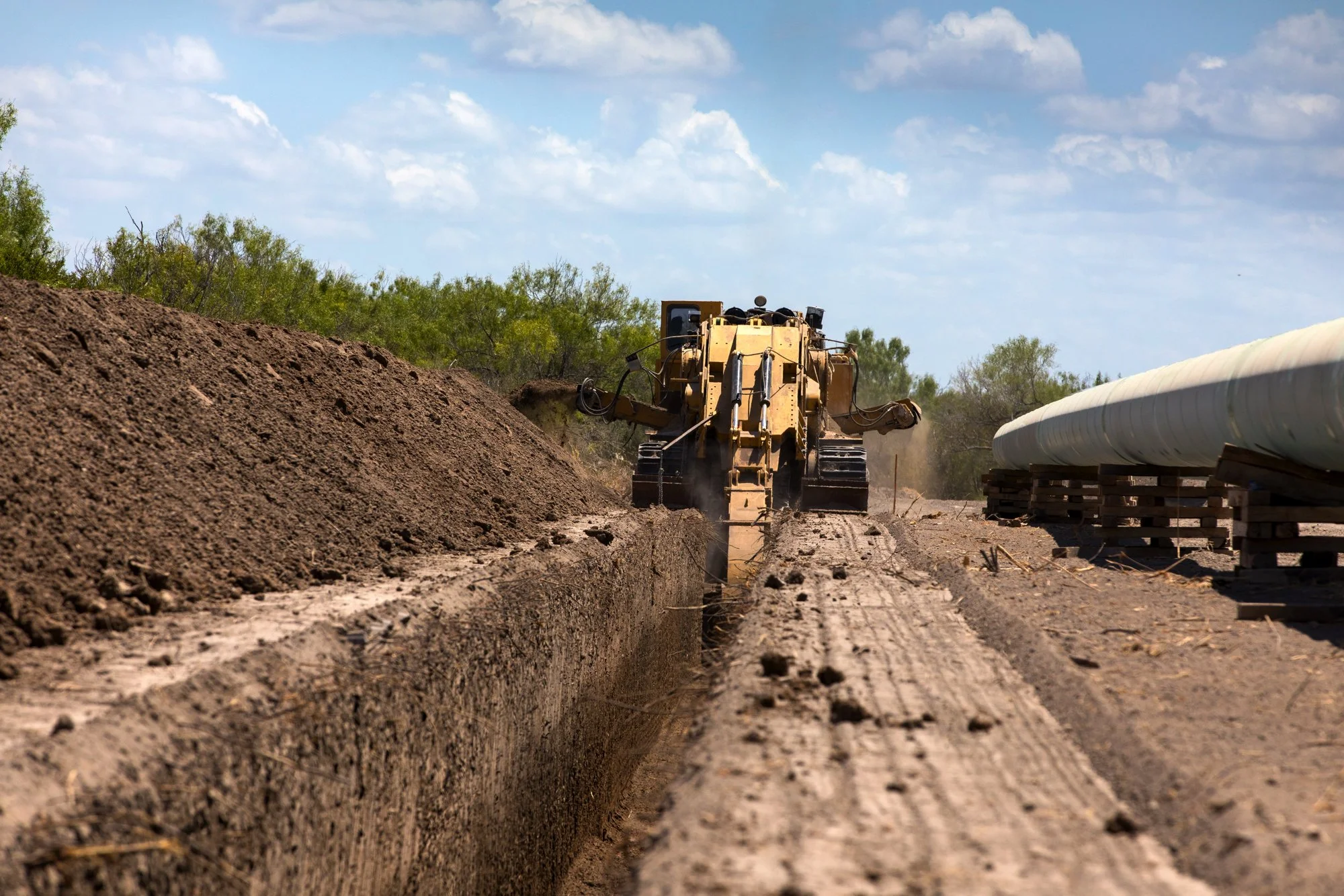 A bulldozer working on a construction site with soil and pipes nearby, under a partly cloudy sky.