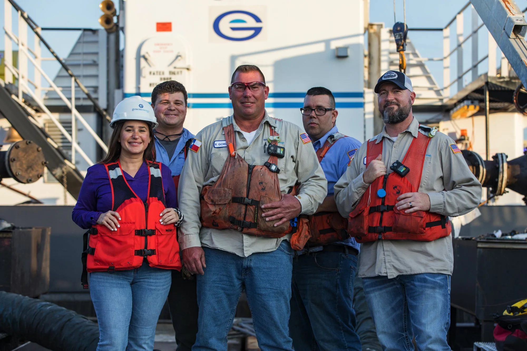 Five people standing together on an industrial site, wearing safety gear and life jackets, smiling at the camera, with a large machinery or equipment in the background.