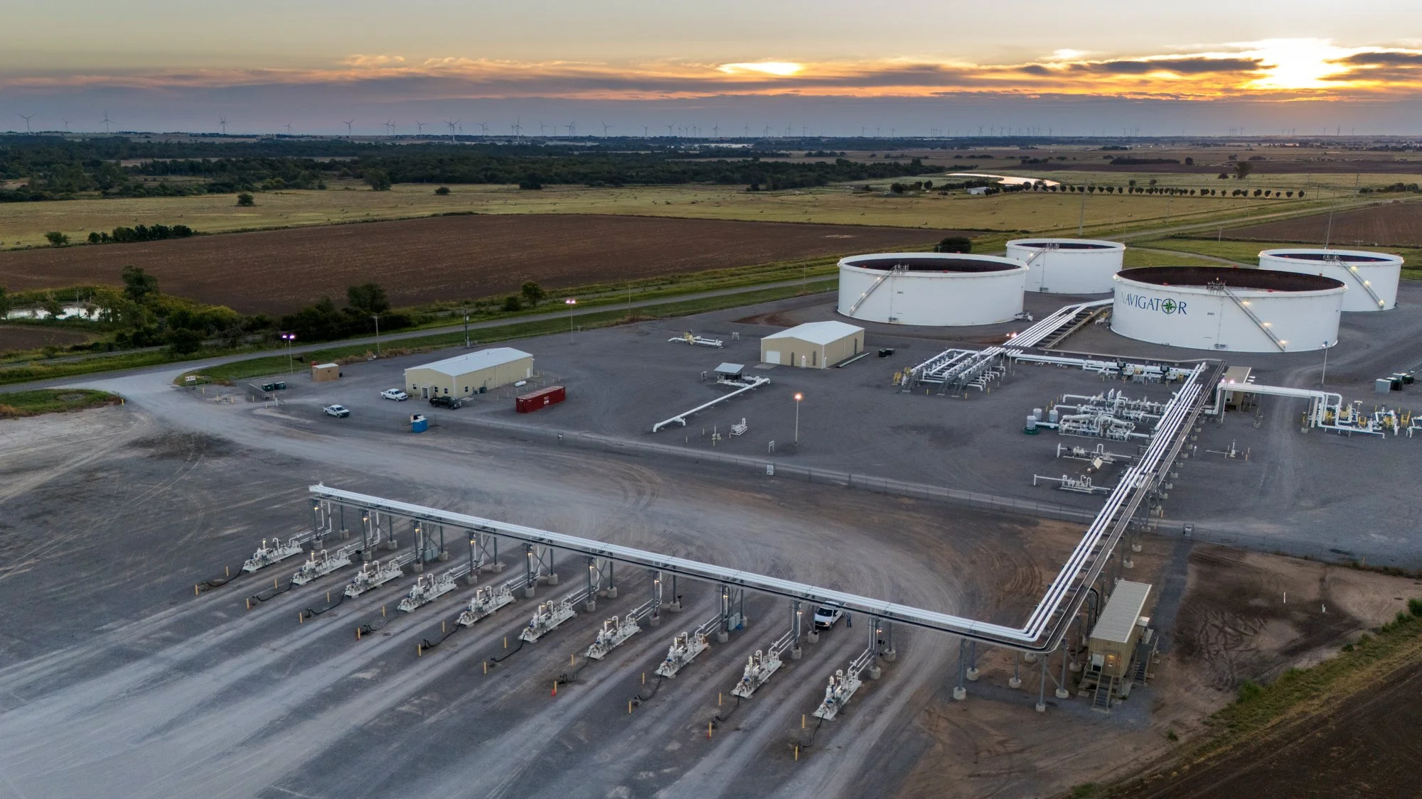 Aerial view of a renewable energy plant with large white storage tanks and pipelines, set against farmland under a sunset sky.
