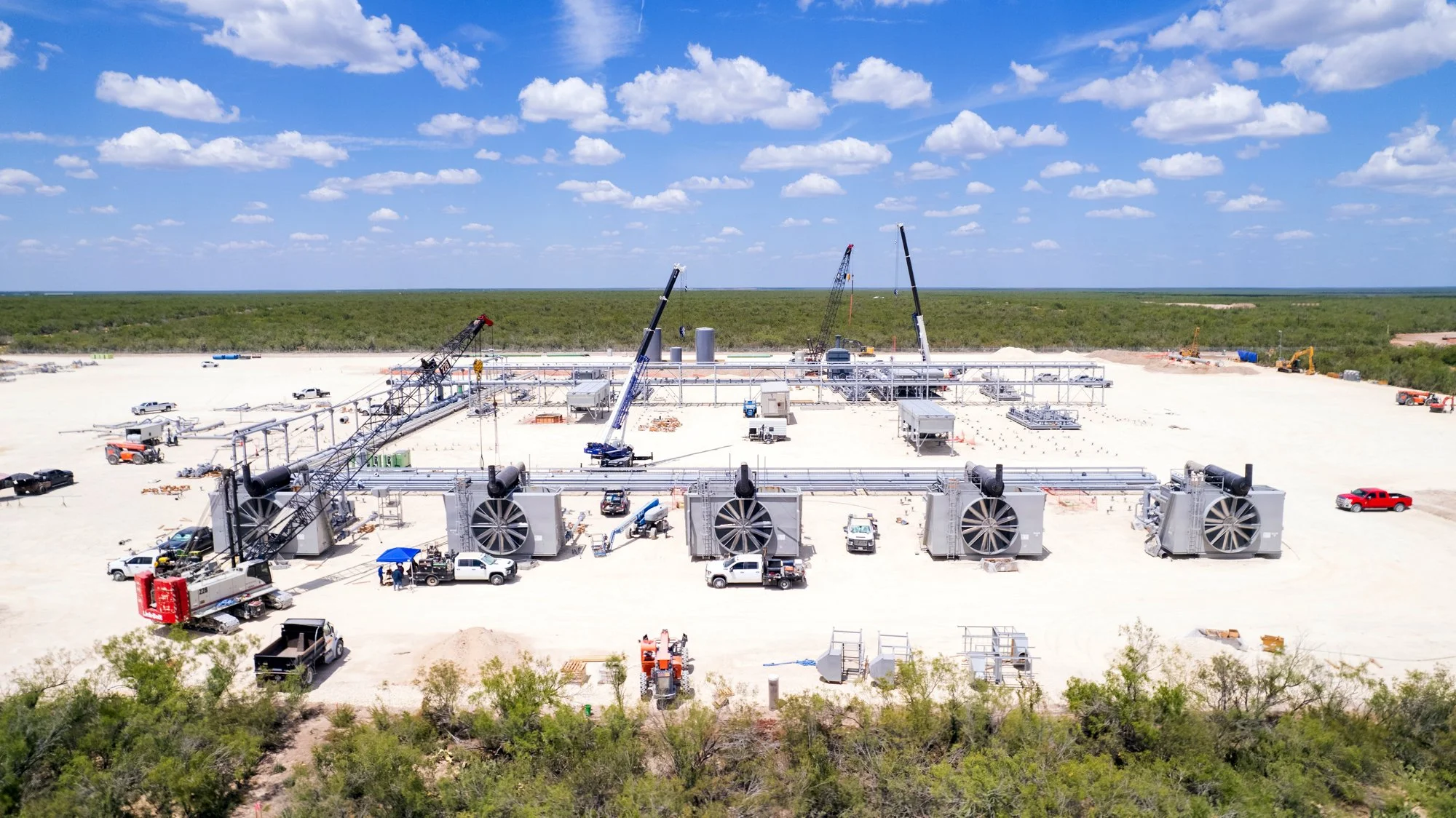 Construction site with large industrial equipment, cranes, trucks, and pipes on a flat, sandy terrain under a blue sky with scattered clouds.