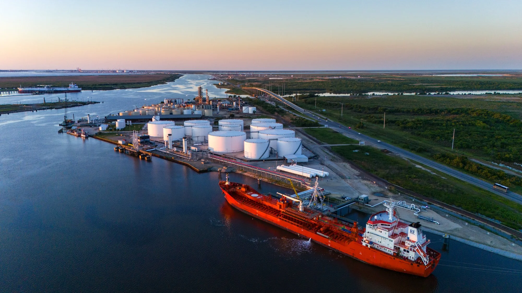 An aerial view of an industrial port area with large white storage tanks, a red cargo ship docked in the water, and a highway running parallel to the coast, with lush green fields and water bodies in the background during sunset.