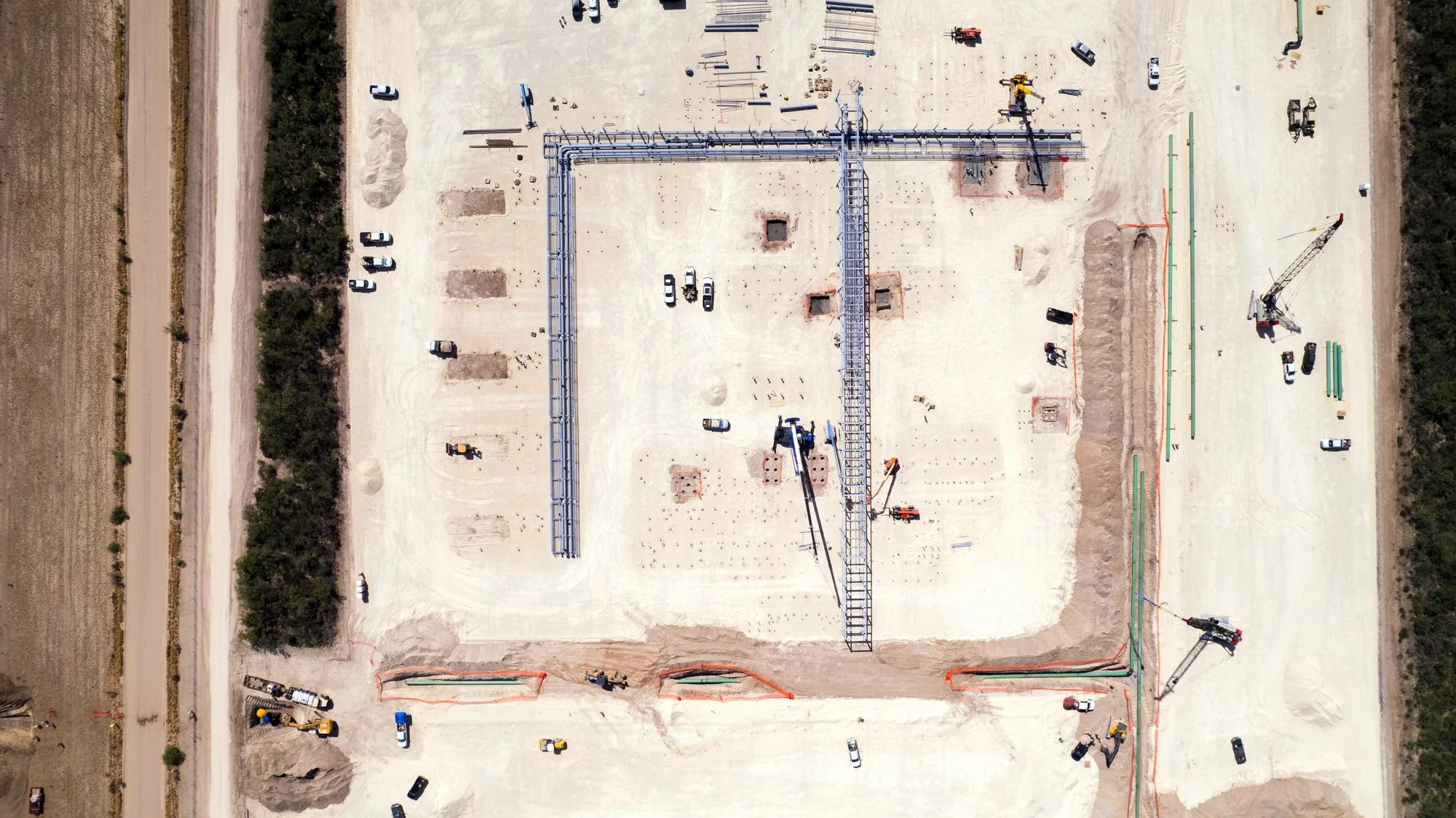 An aerial view of a construction site with workers, vehicles, and structural frameworks on a sandy area, surrounded by dirt roads and greenery.