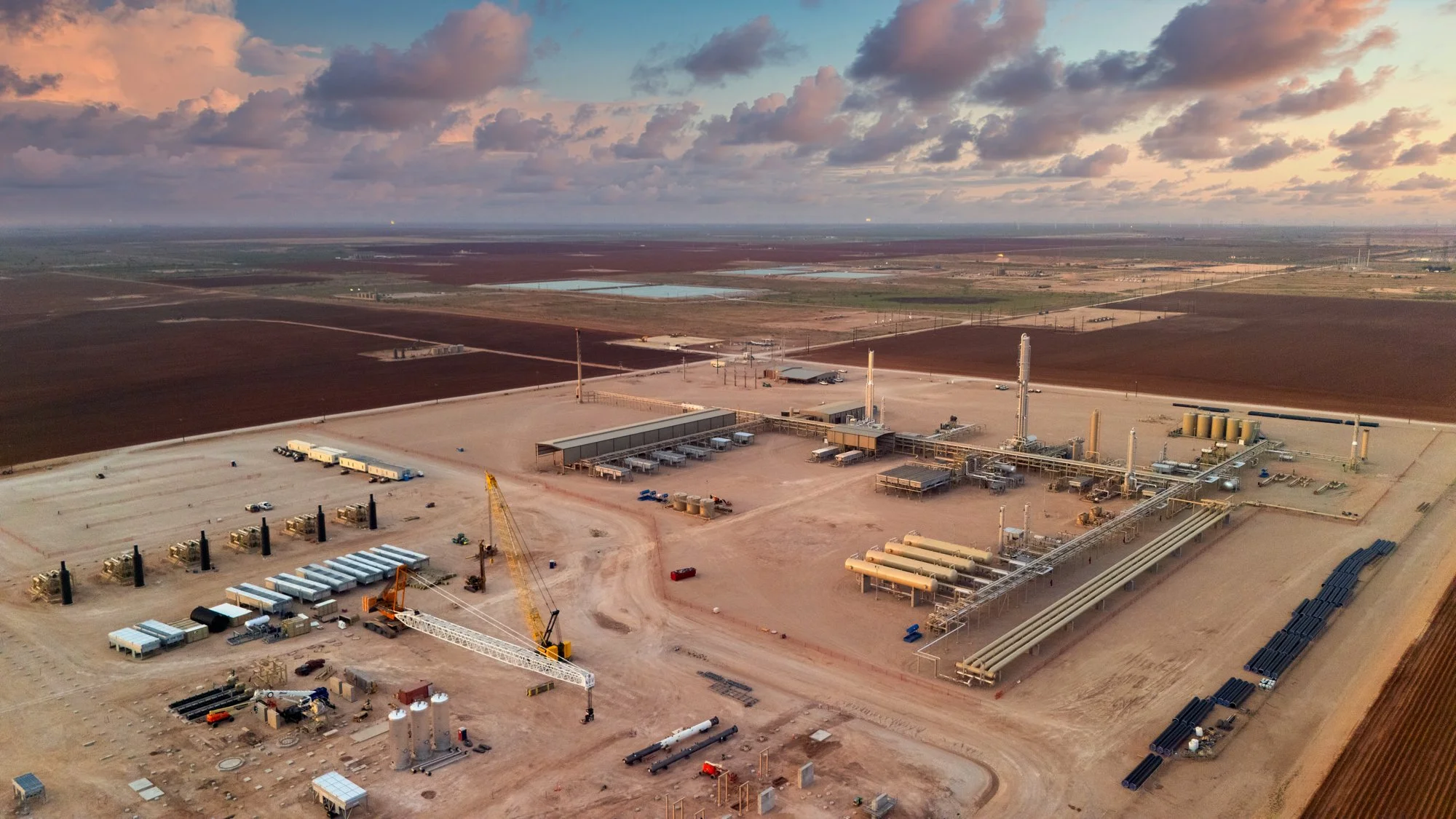 An aerial view of an industrial site under construction with pipelines, cranes, and storage tanks, located in a flat, rural landscape with fields and ponds in the background, under a cloudy sky.