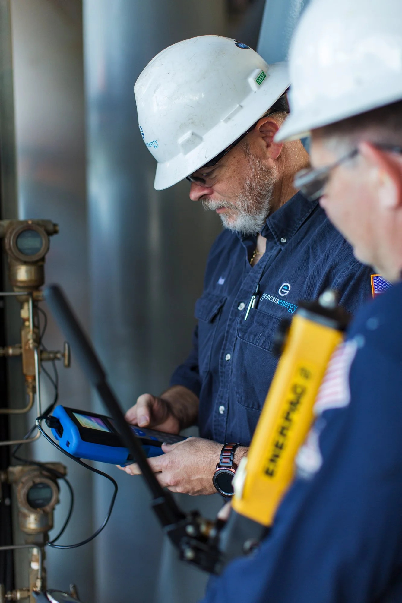 Two energy workers in blue uniforms and white safety helmets inspecting equipment and using diagnostic tools.