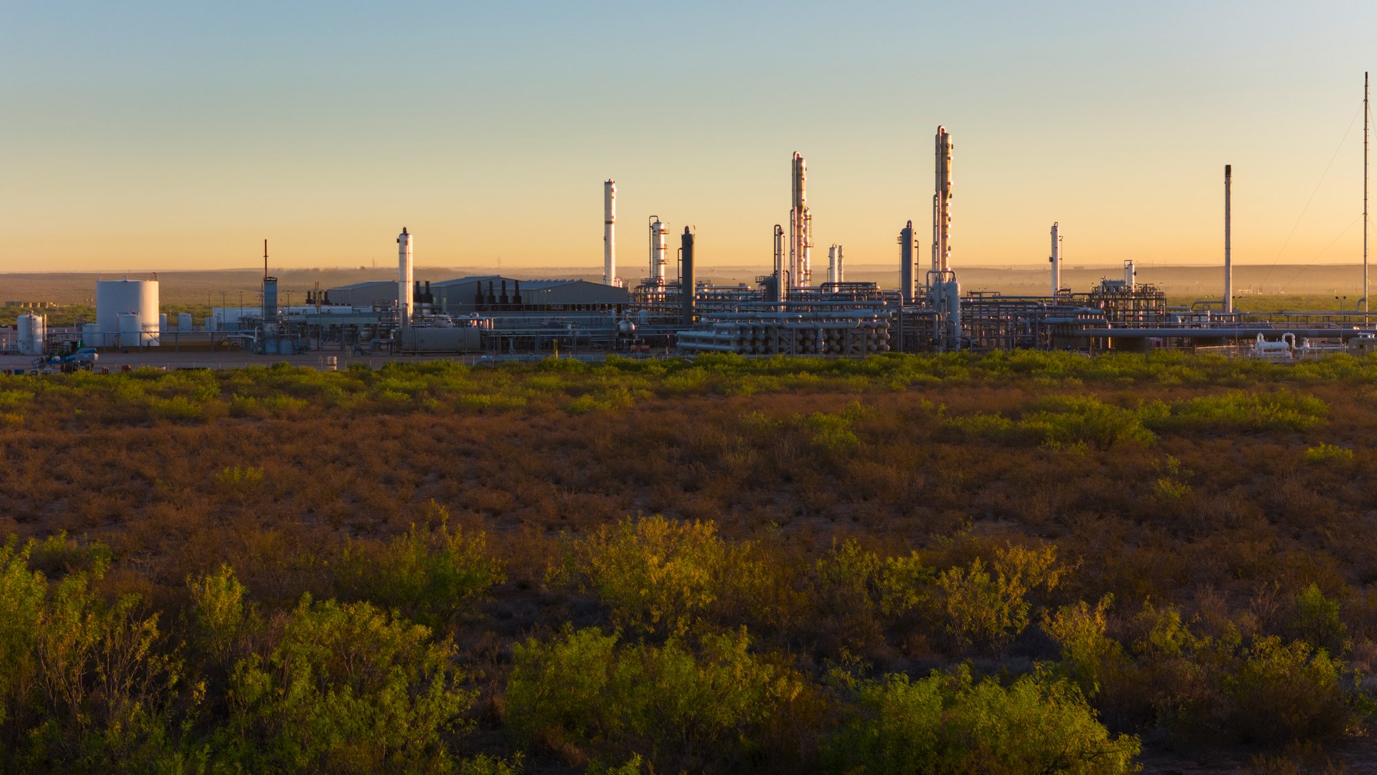 An industrial oil refinery with multiple tall chimneys and processing units, set against a backdrop of a clear sky during sunset, with green and brown desert vegetation in the foreground.