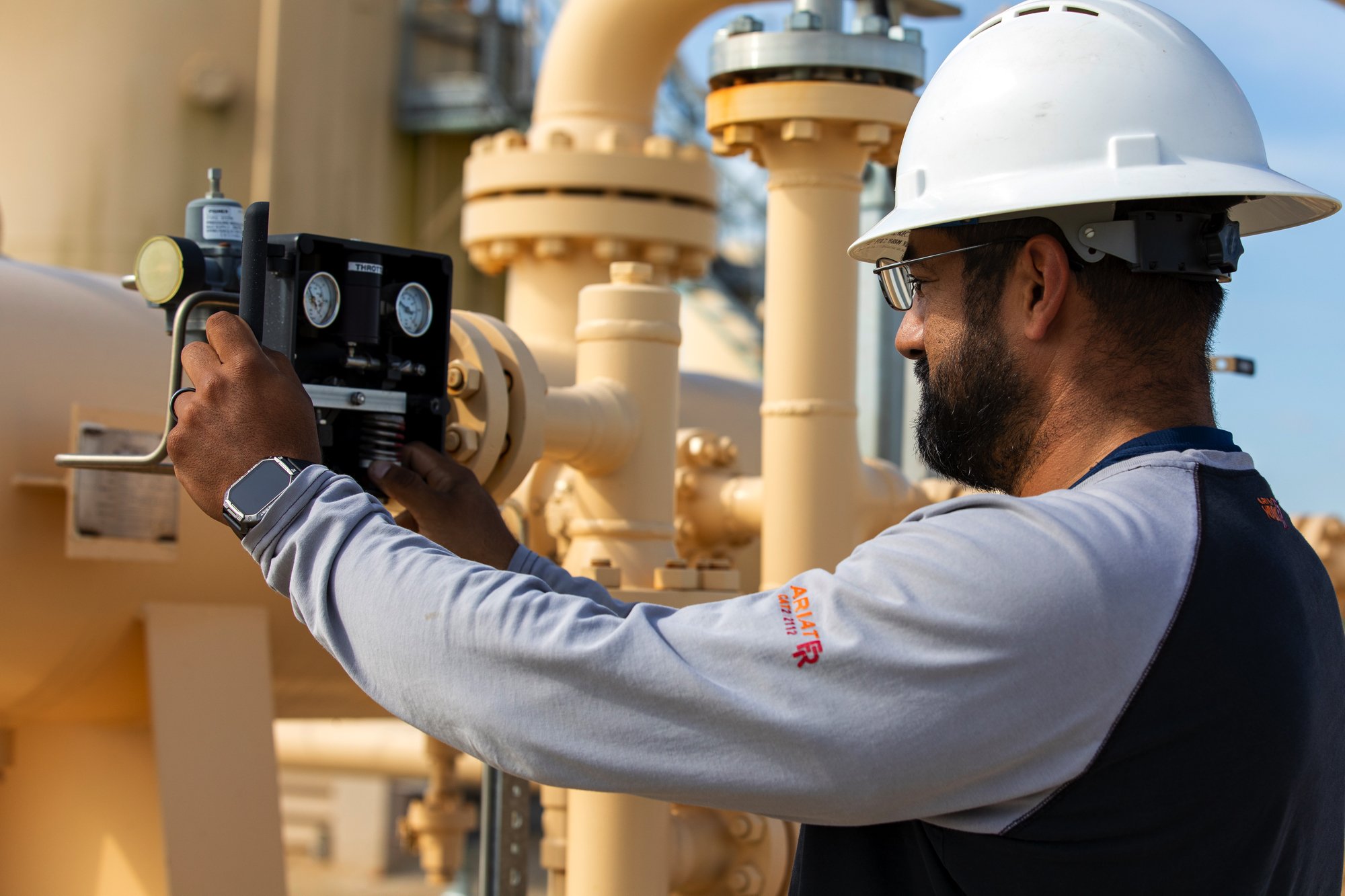 A man with a beard wearing safety glasses and a white helmet working with industrial equipment. He is adjusting or inspecting a valve or control device on a yellow pipeline system outdoors.