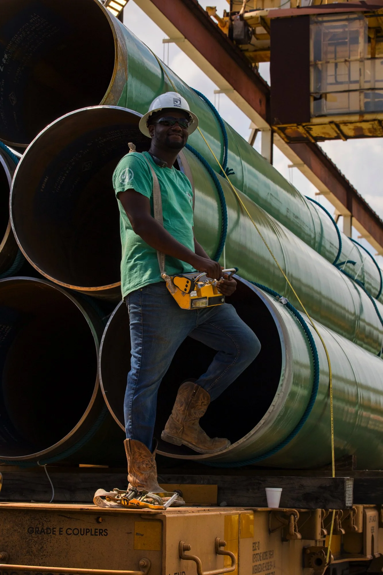 A worker standing on a flatbed truck among large green pipes at an industrial site, wearing a hard hat and safety glasses, holding a yellow chainsaw.