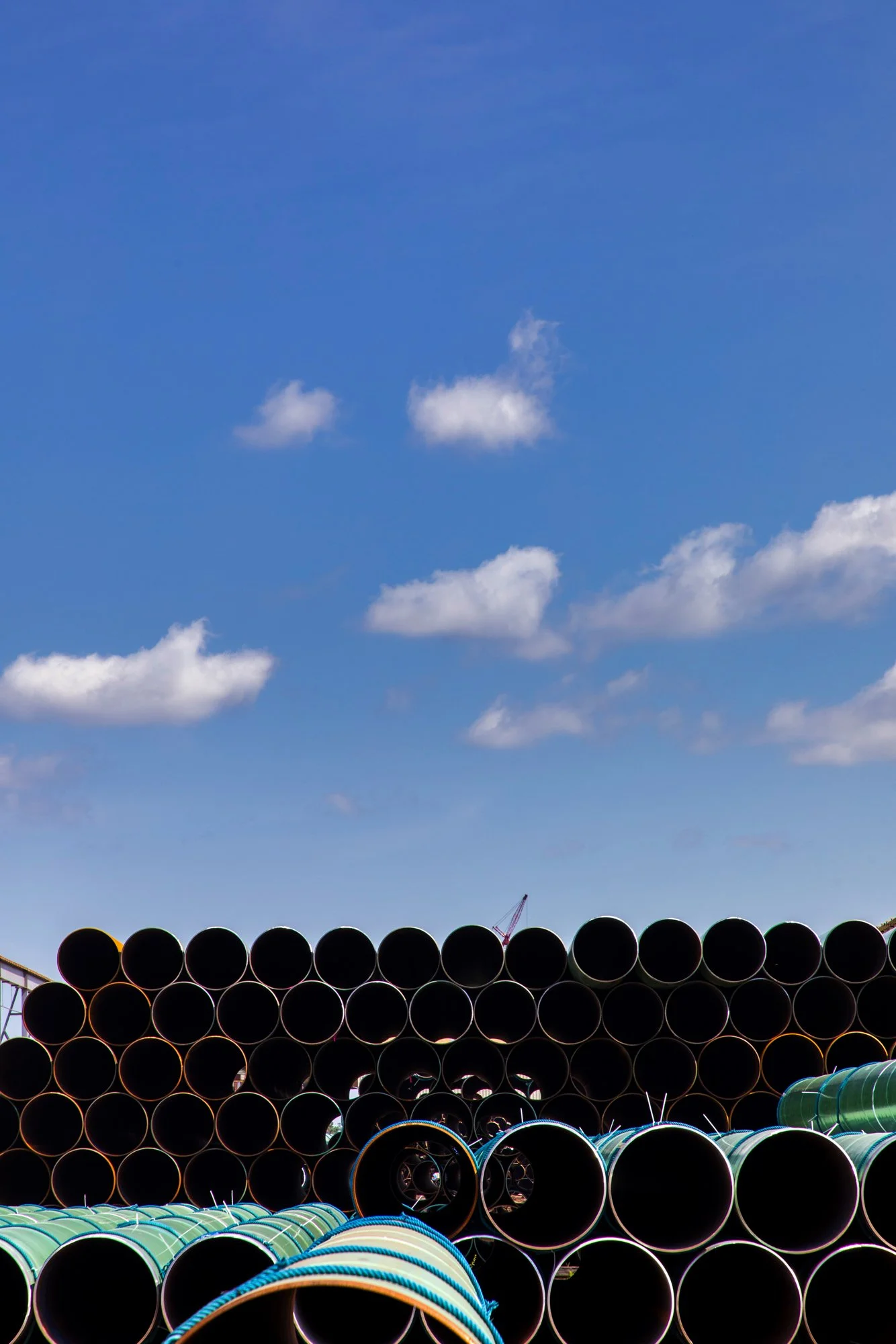 Stacked black and green pipes at a construction site under a blue sky with scattered clouds.