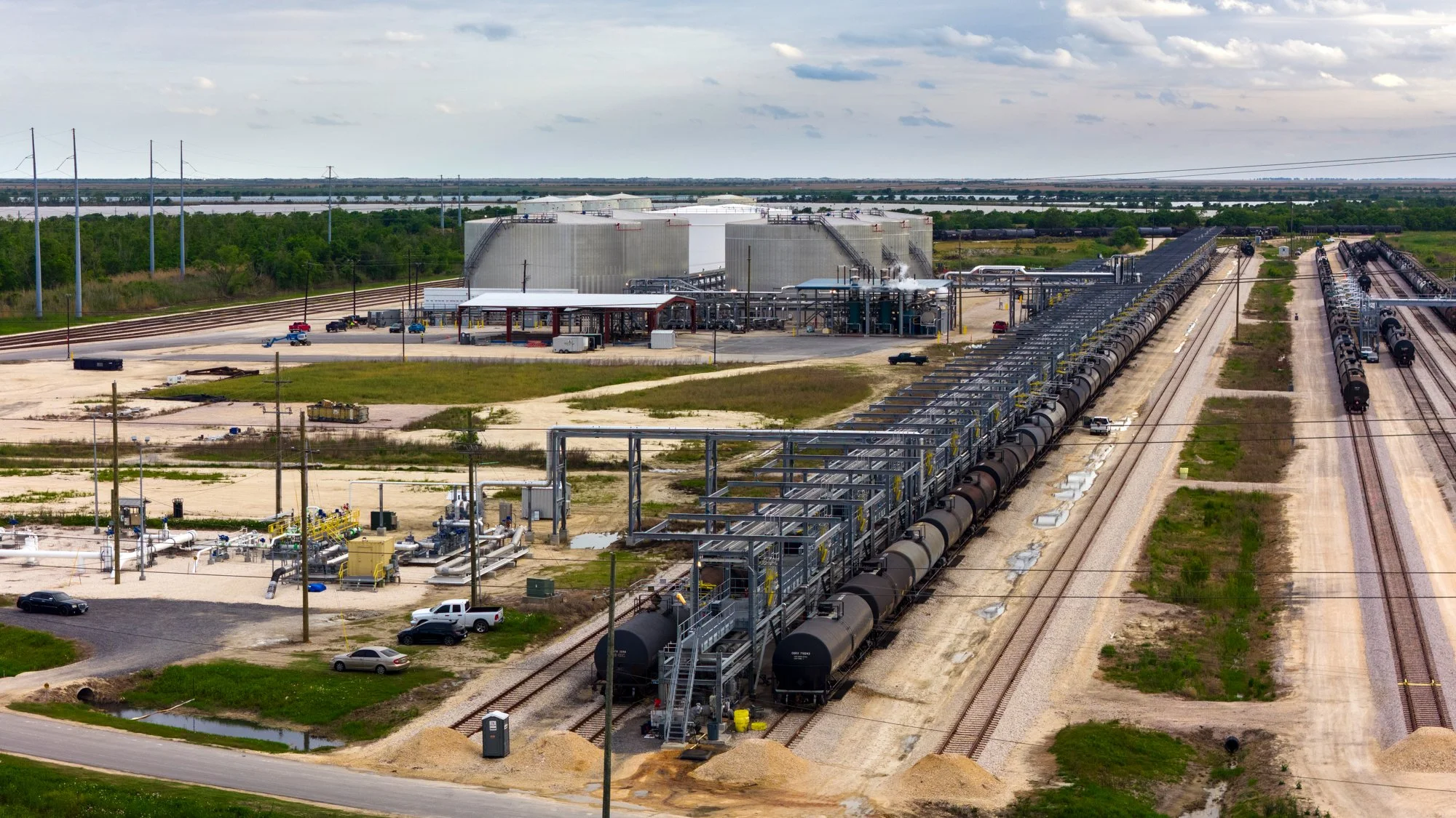 An industrial facility with multiple train tracks, tanker cars, and large storage tanks in a rural area with greenery and overcast sky.