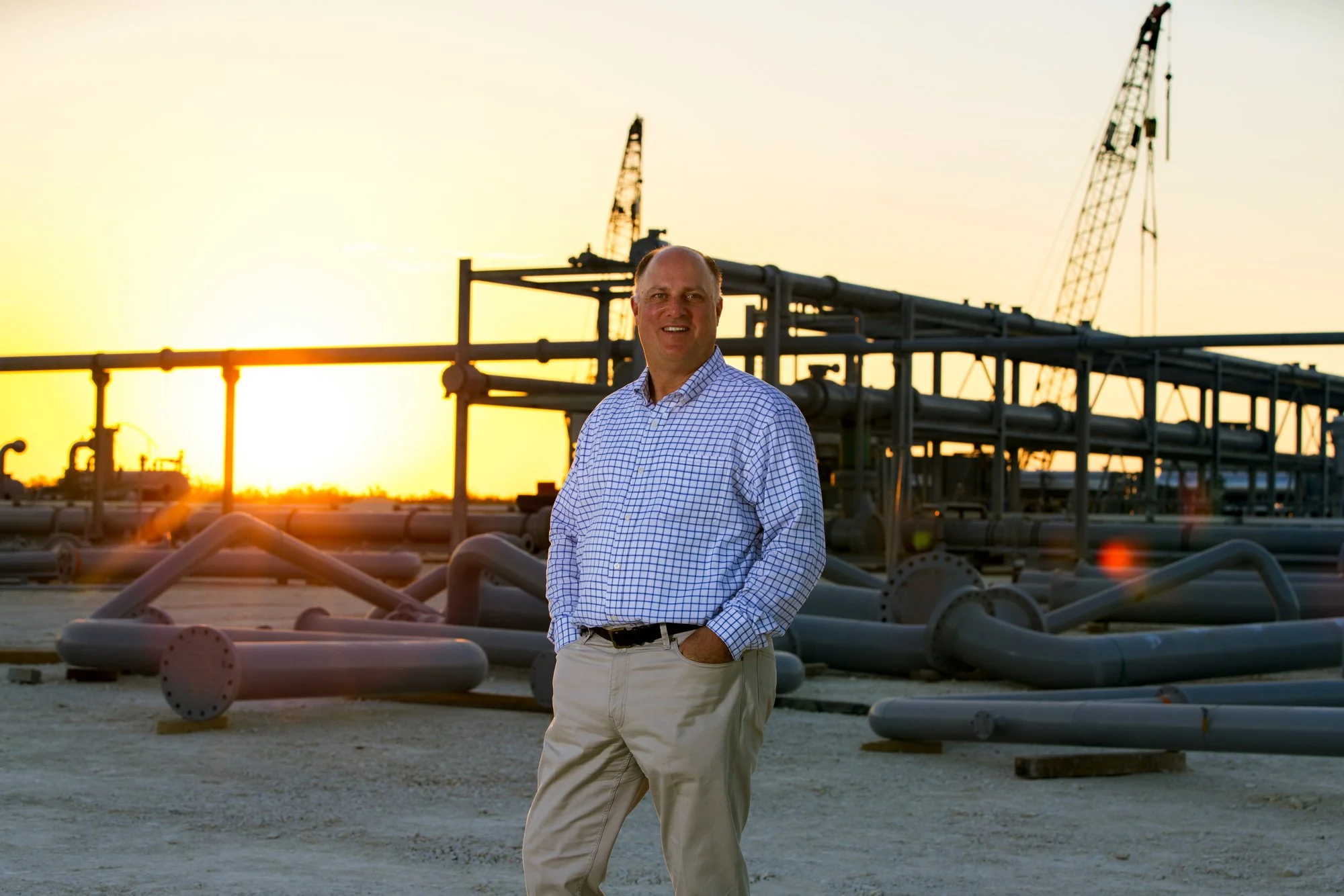 A man in a checkered shirt and khakis stands in front of an oil or gas pipeline infrastructure at sunset.