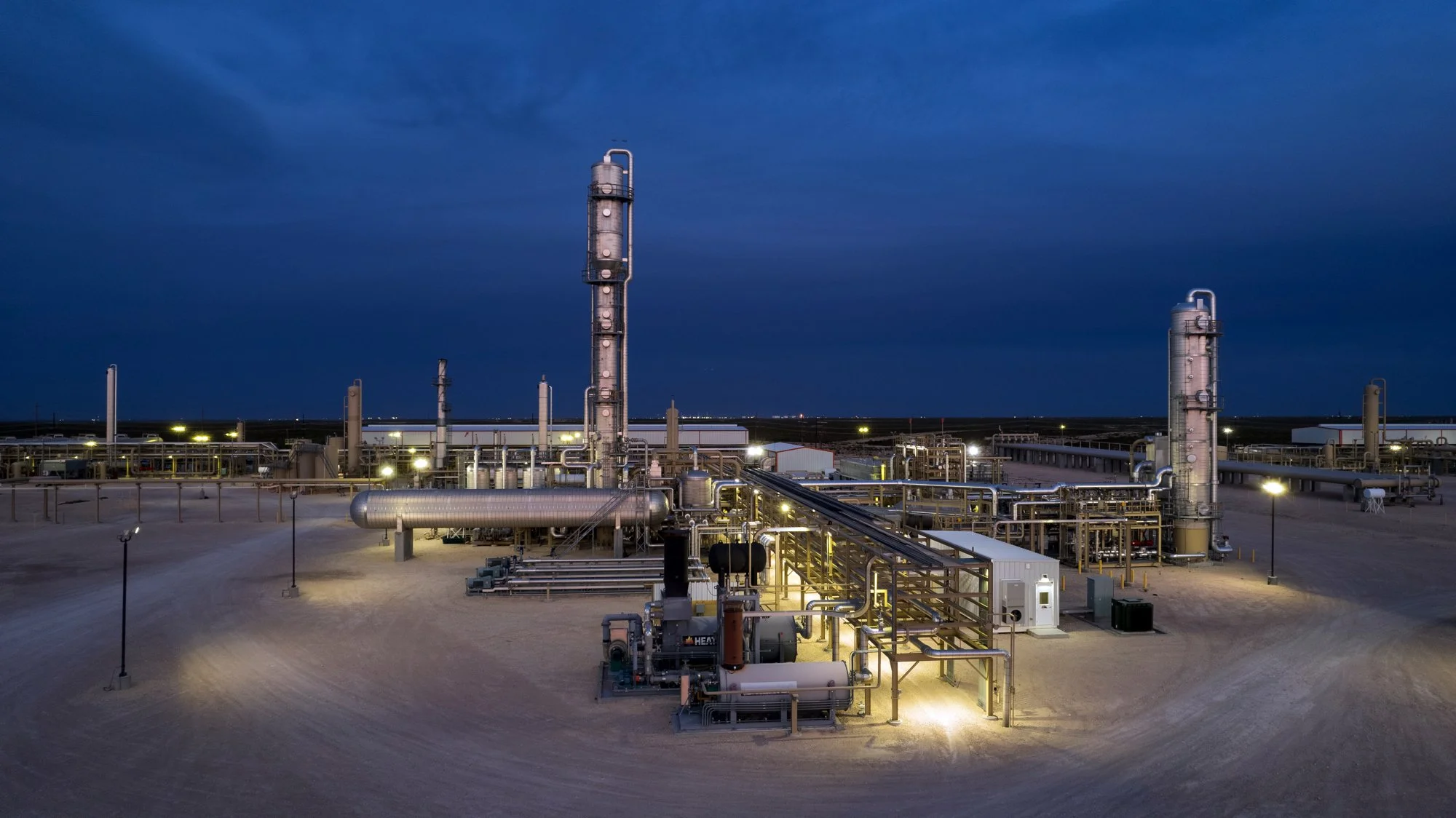 An industrial oil or gas processing facility at night with illuminated pipelines, towers, and equipment against a dark sky.