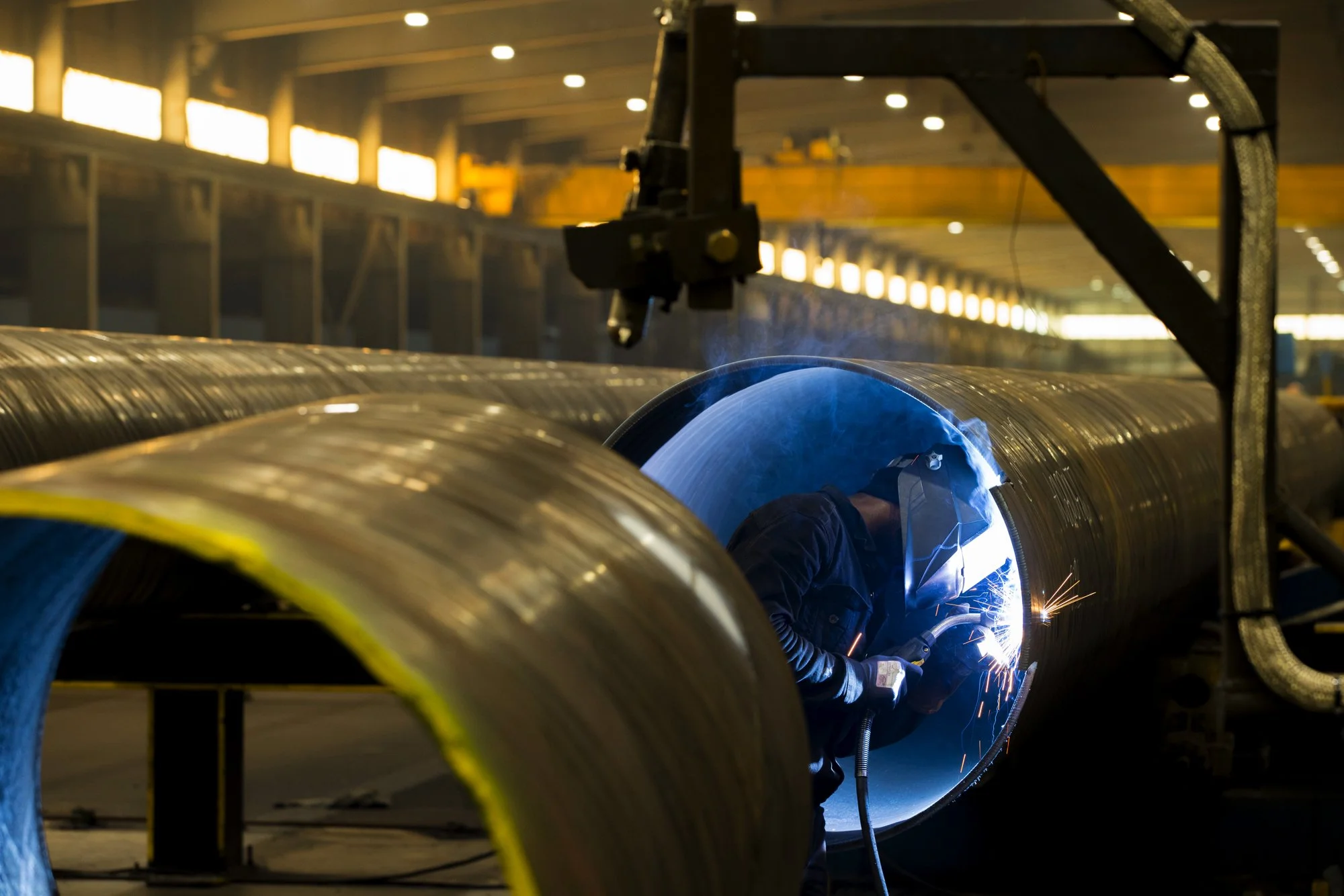 A worker welding inside a large metal pipe in an industrial warehouse.