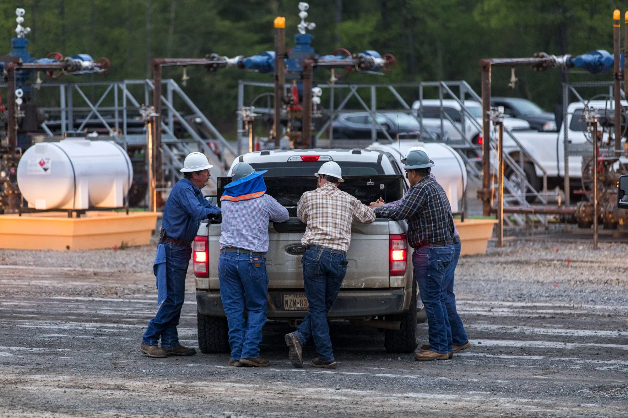 Five workers wearing safety helmets and work clothes gather around the back of a pickup truck at an industrial site with pipelines and equipment in the background.