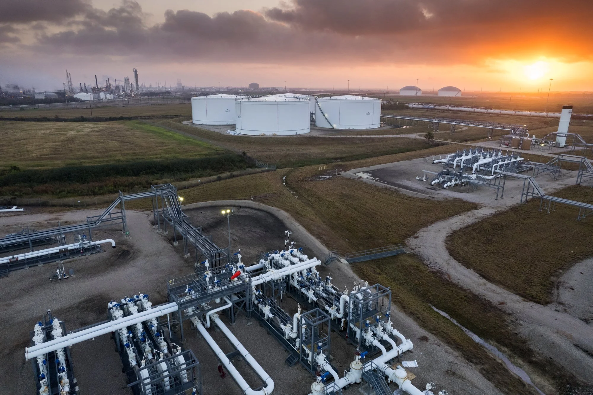 Aerial view of an oil storage facility with large white tanks, pipelines, and industrial structures at sunset.