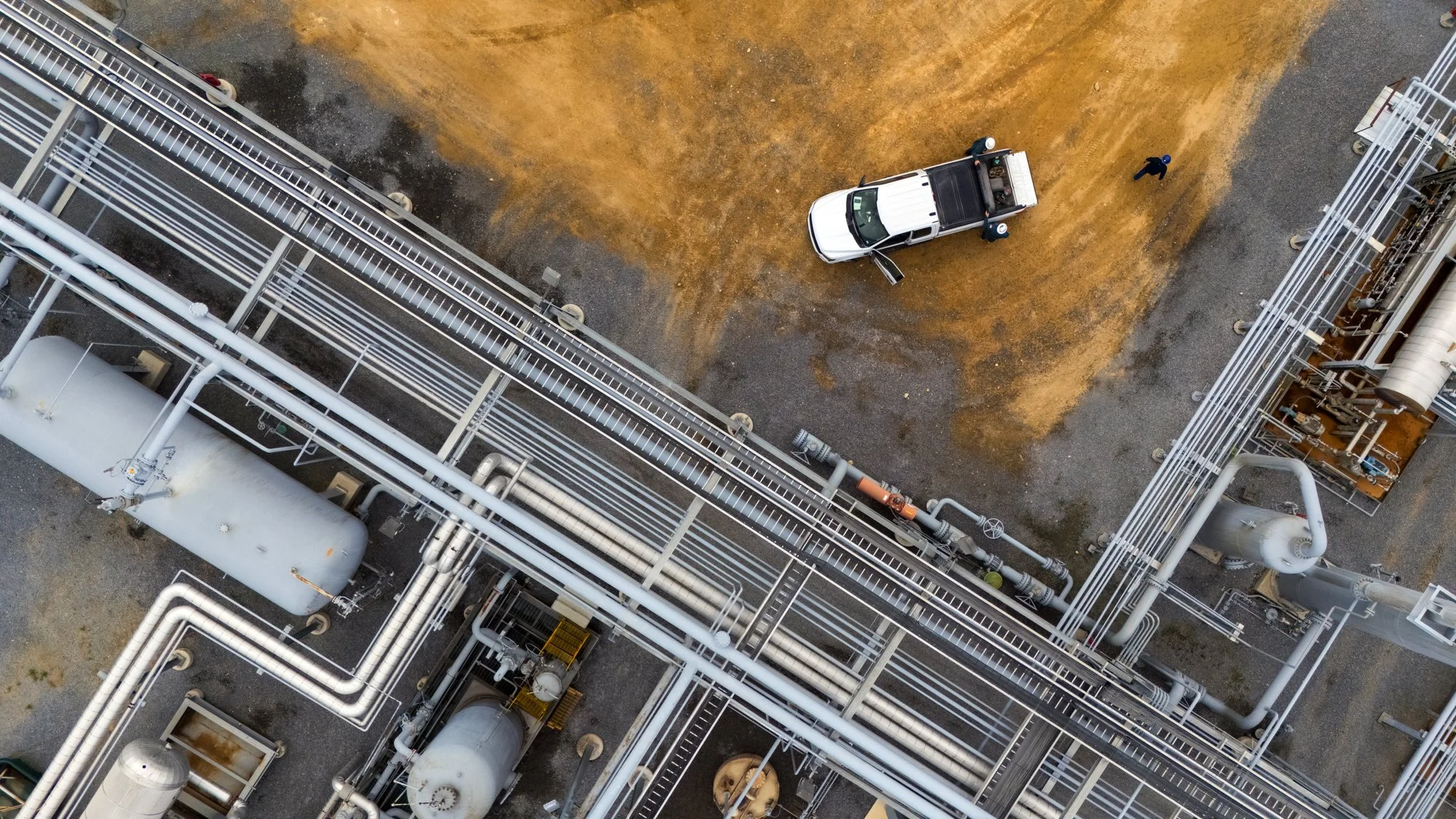 An aerial view of an industrial facility with metal pipes and tanks, a white pickup truck on dirt, and two workers near the truck.
