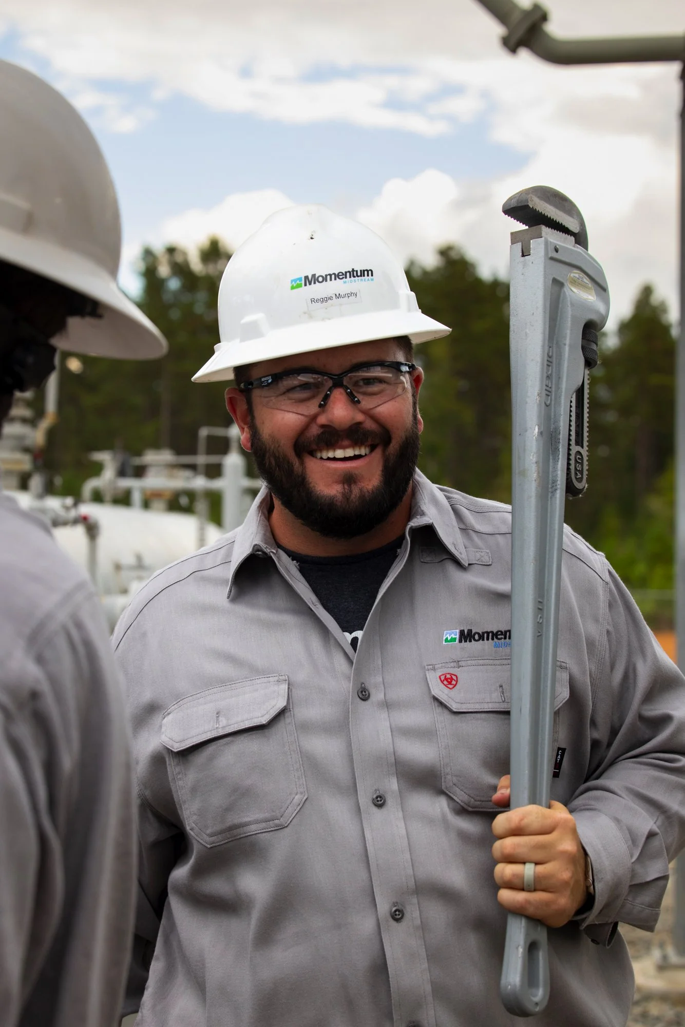 A smiling man in safety glasses, a white hard hat, and a grey work shirt holding a pipe wrench while standing outdoors at a construction site, with trees and equipment in the background.