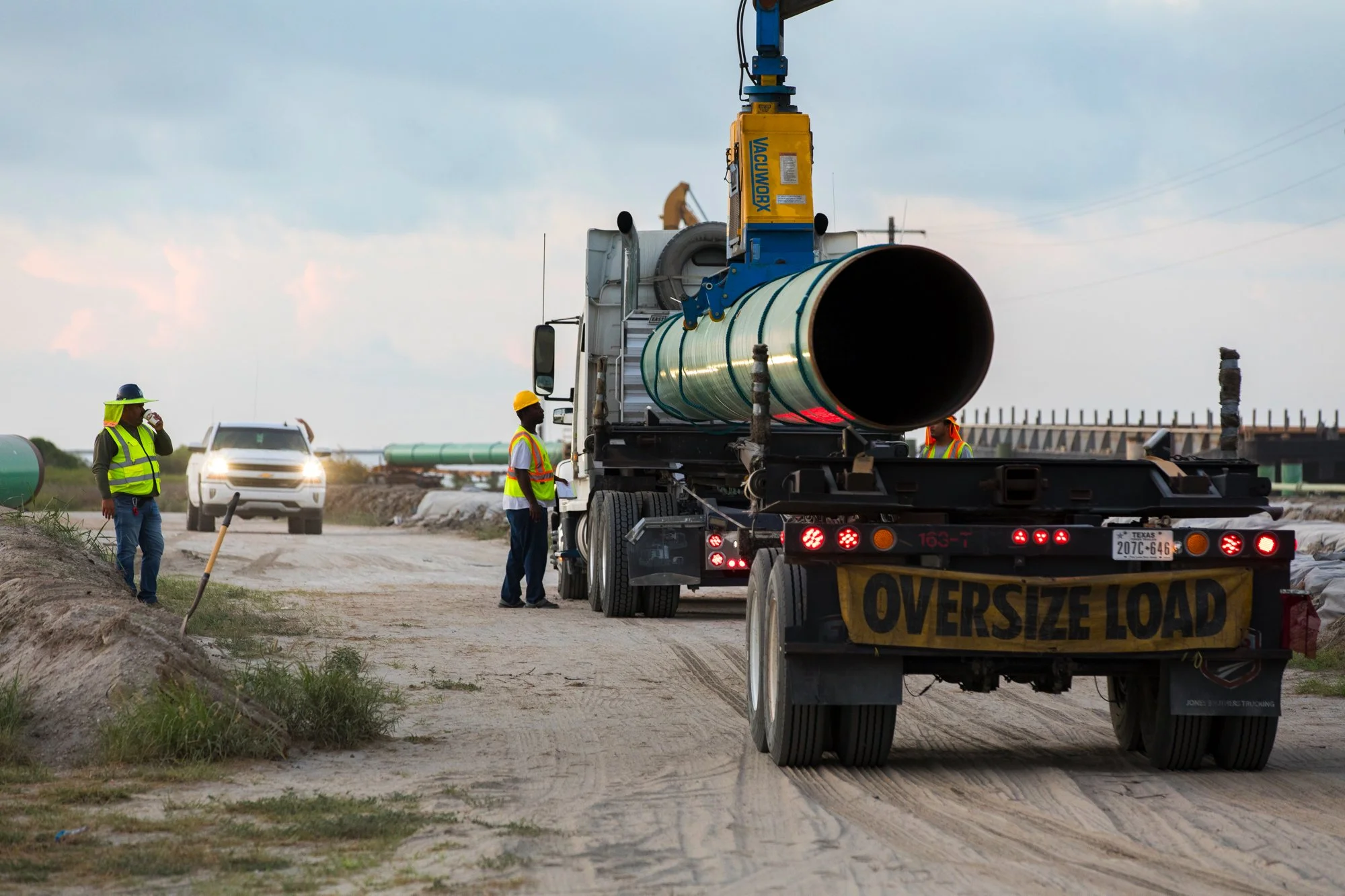 A construction site with workers wearing safety vests and helmets, a large truck with an oversize load sign, and an oversized pipe being installed or transported.