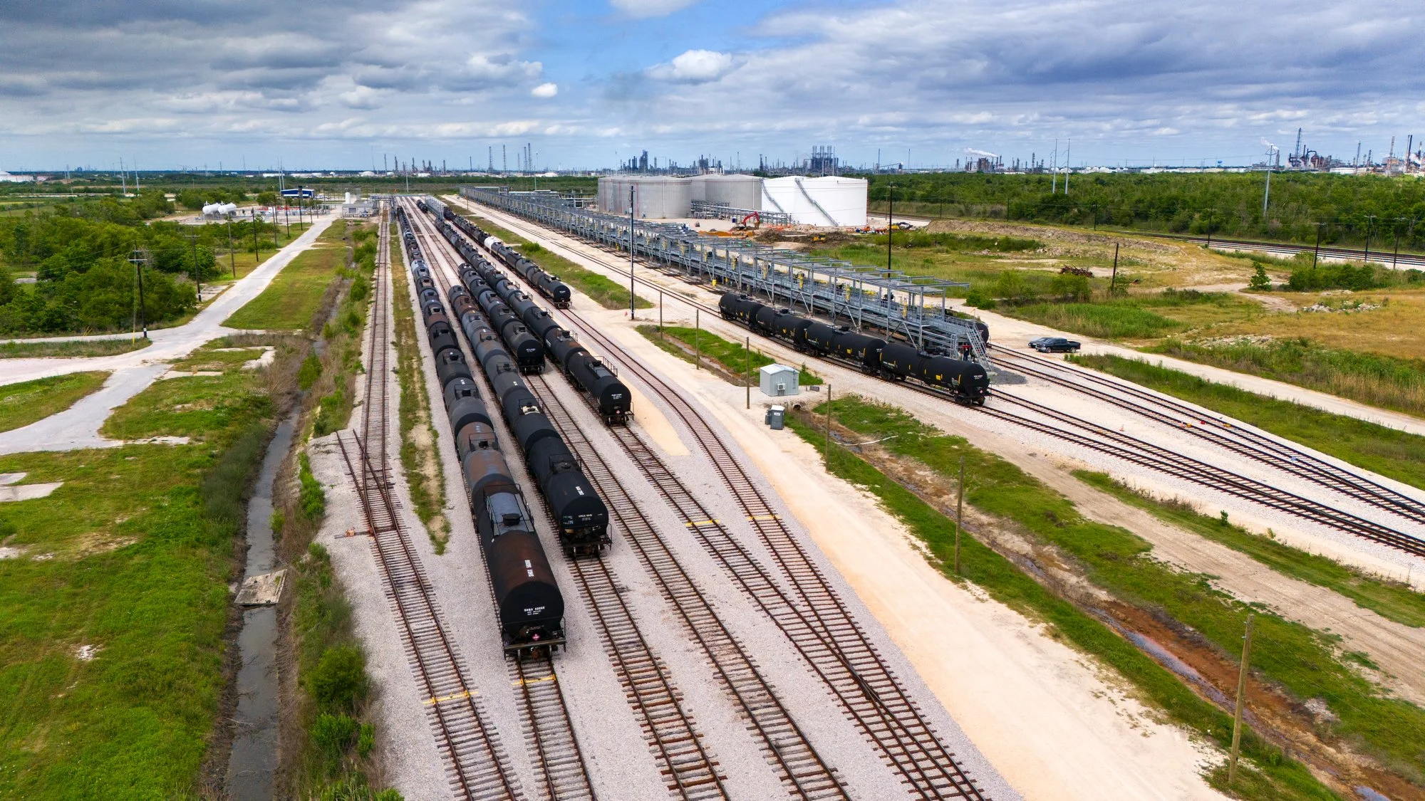 Multiple freight train cars, including black tankers and boxcars, on railroad tracks in an industrial area with storage tanks and factories in the background.