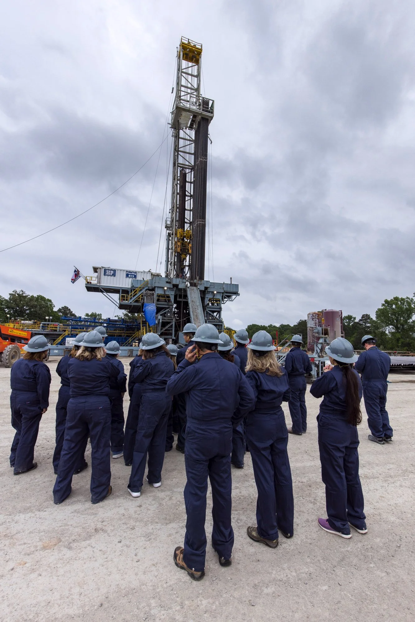 A group of people wearing navy blue coveralls and gray hard hats gathered outdoors at an oil drilling site, looking at a large drilling rig against a cloudy sky.