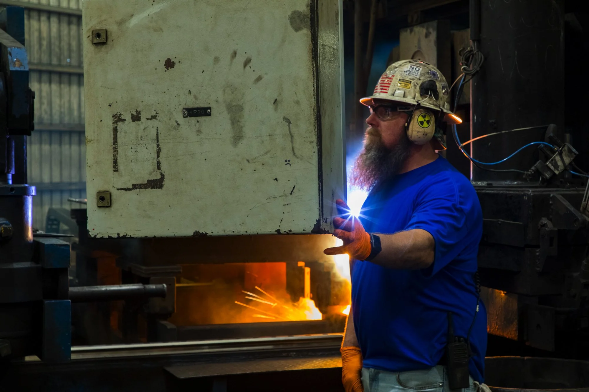 A man welding in an industrial workshop, wearing safety gear including a helmet with American flag and radiation symbols, goggles, gloves, and a blue shirt, with sparks and bright welding light.