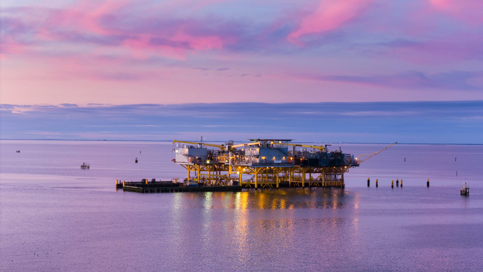 An offshore oil rig illuminated by lights stands in calm water during sunset, with pink and purple clouds in the sky.